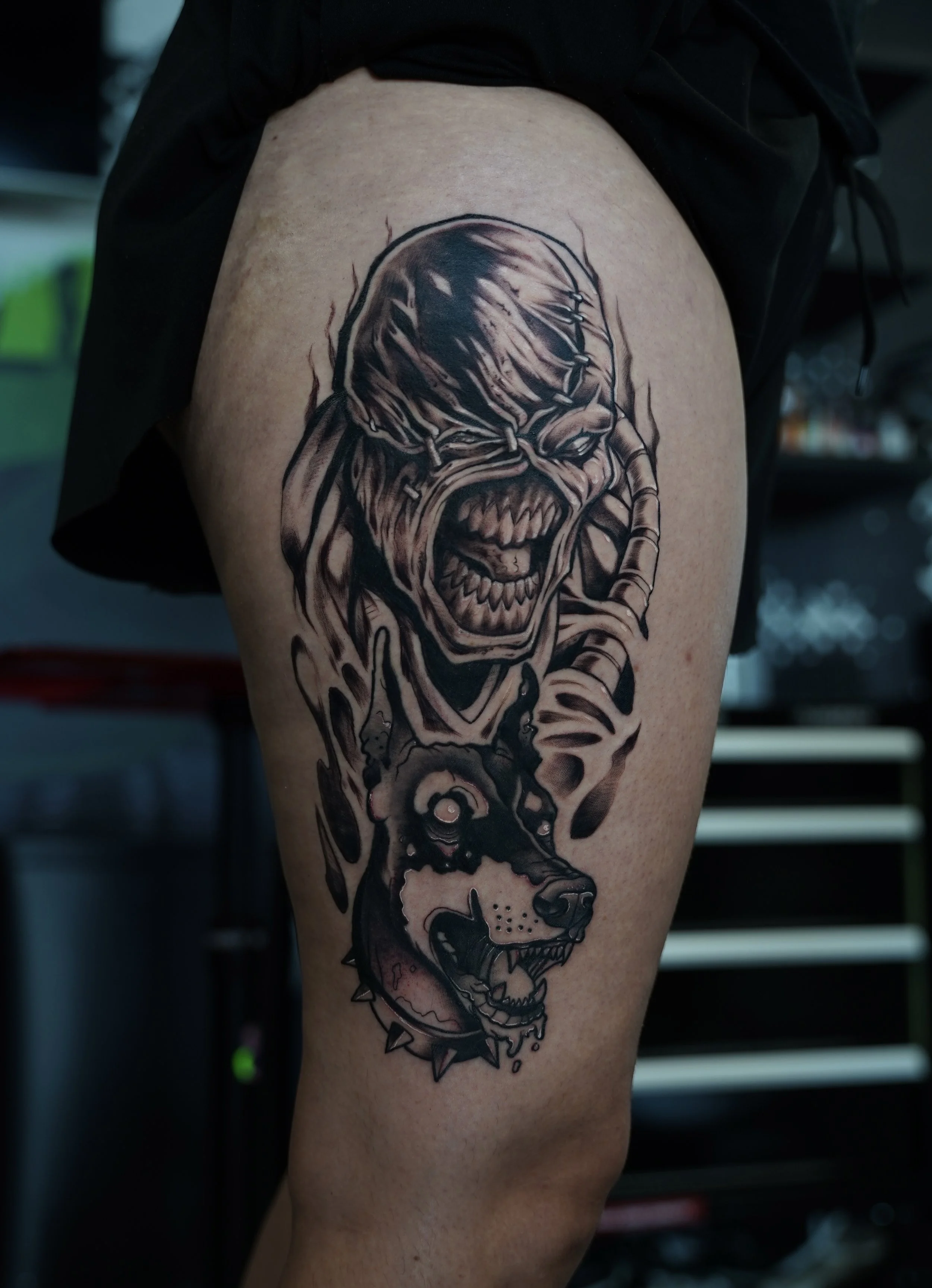 A tattoo on a person's thigh featuring the face of a snarling, zombie-like creature and the head of a snarling dog, both in black and gray ink with detailed shading.