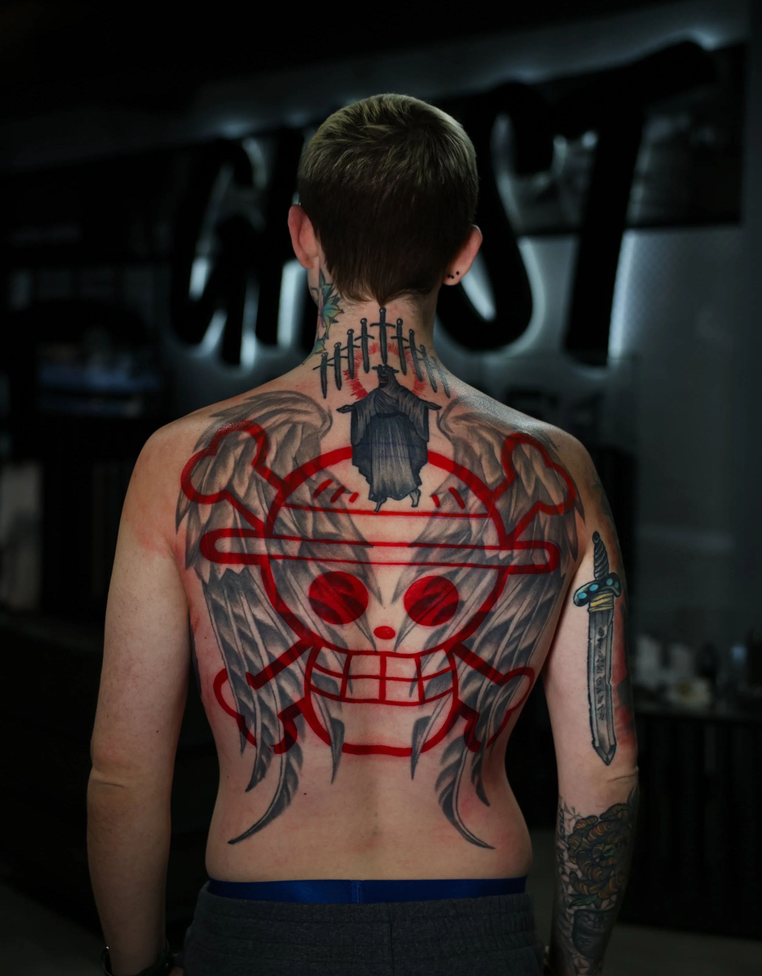 Back of a person with tattoos, including a large, colorful design of a smiling doll face with a red and black theme on their back, a sword tattoo on their right arm, and a tattoo of knives and a figure in dark clothing on their neck.