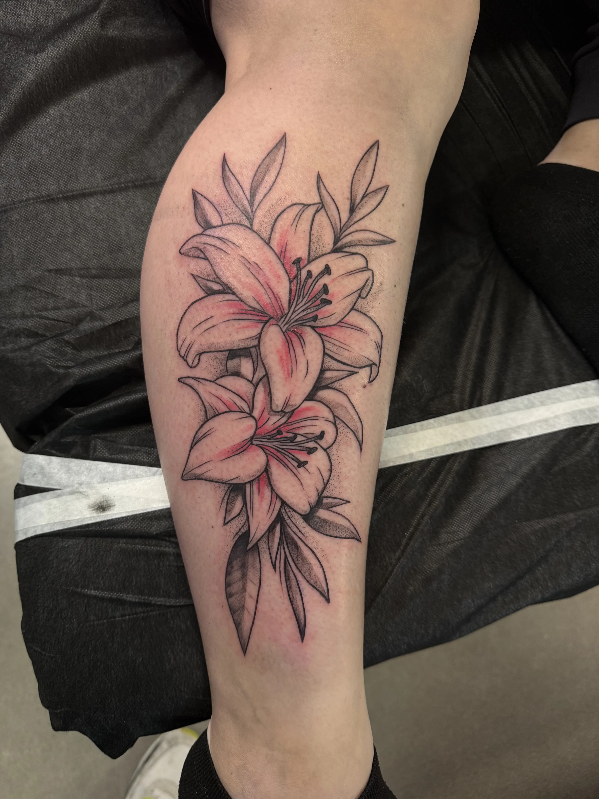 Tattoo of pink lilies with black outlines and green leaves on a person's lower leg.
