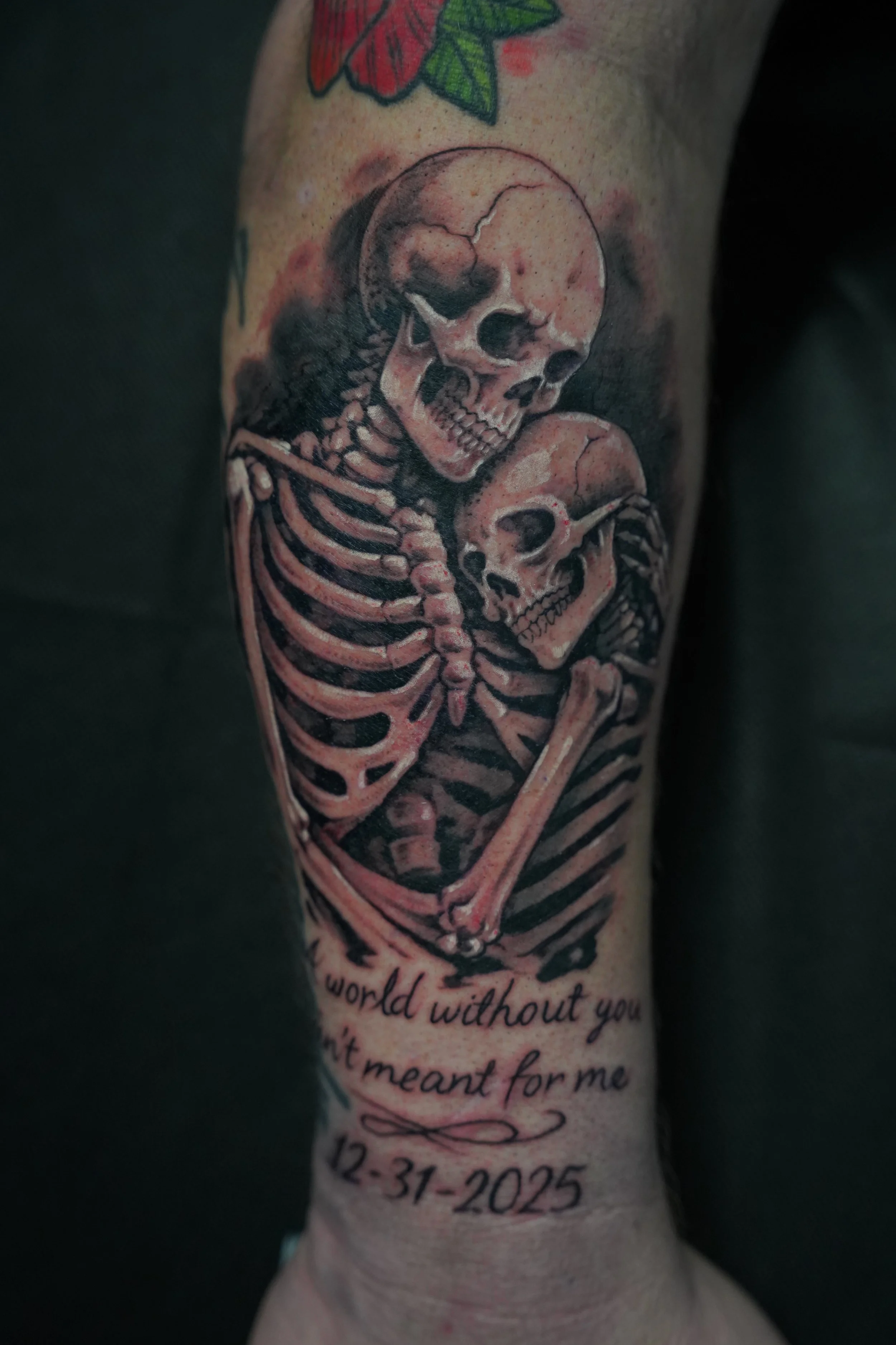 Tattoo of two skeletons embracing with a quote, date, and a small flower above.
