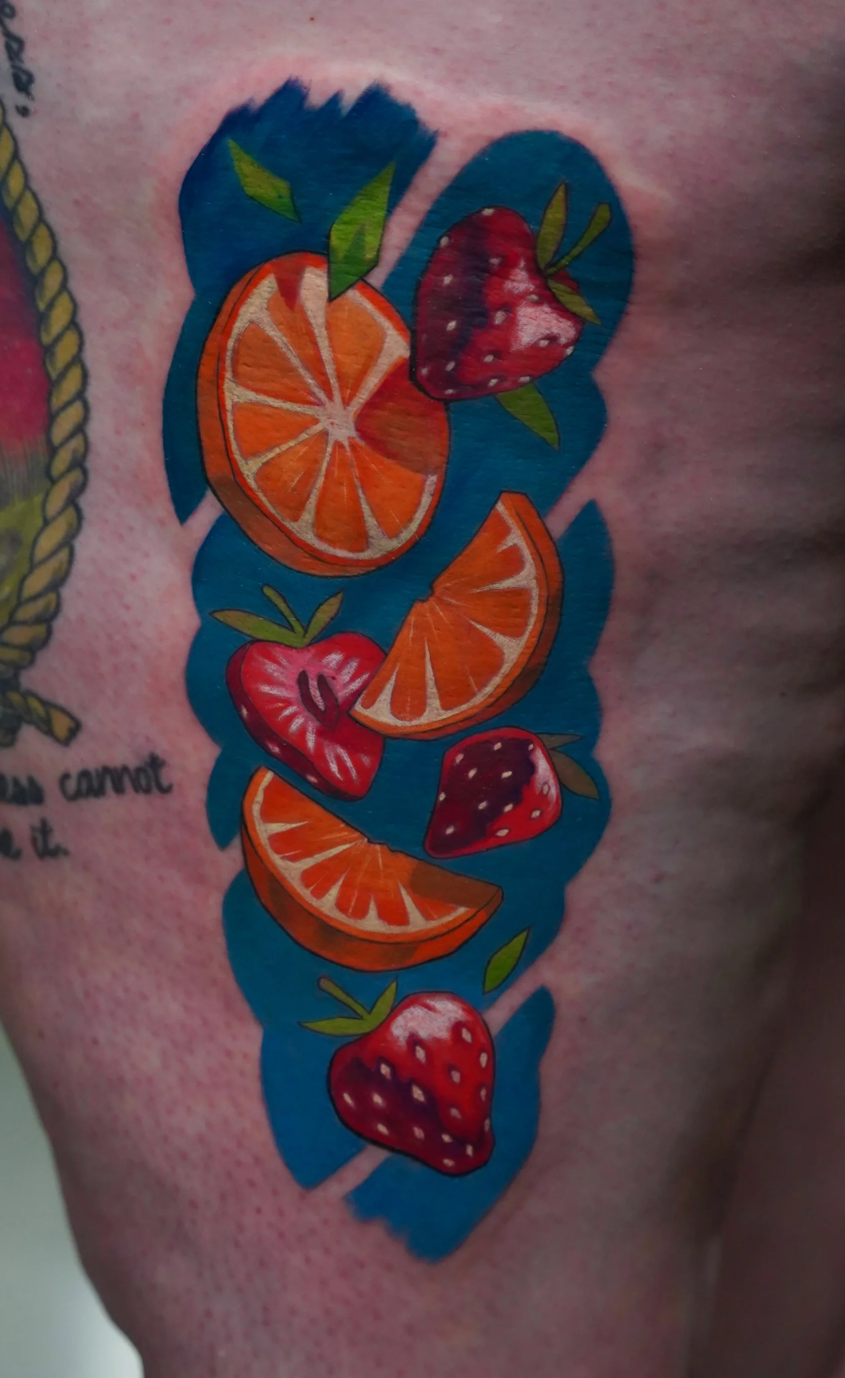 Colorful tattoo of strawberries and oranges on skin with a blue background and some partial black text.