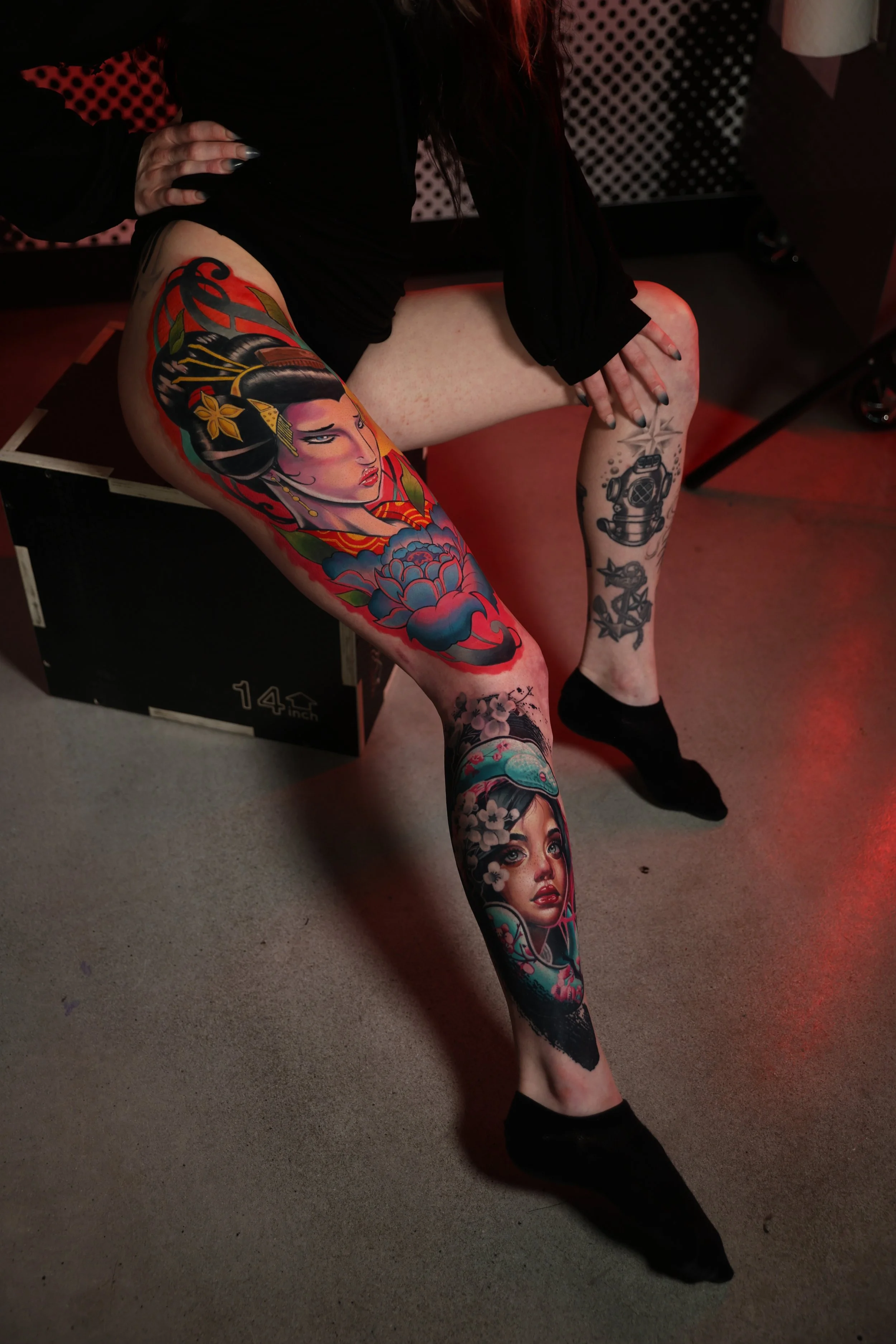 A person with colorful tattoos on both legs, one featuring a woman with traditional Japanese hair and attire, and the other with a woman with flowers in her hair, sitting on a black box in a dimly lit room with red lighting.