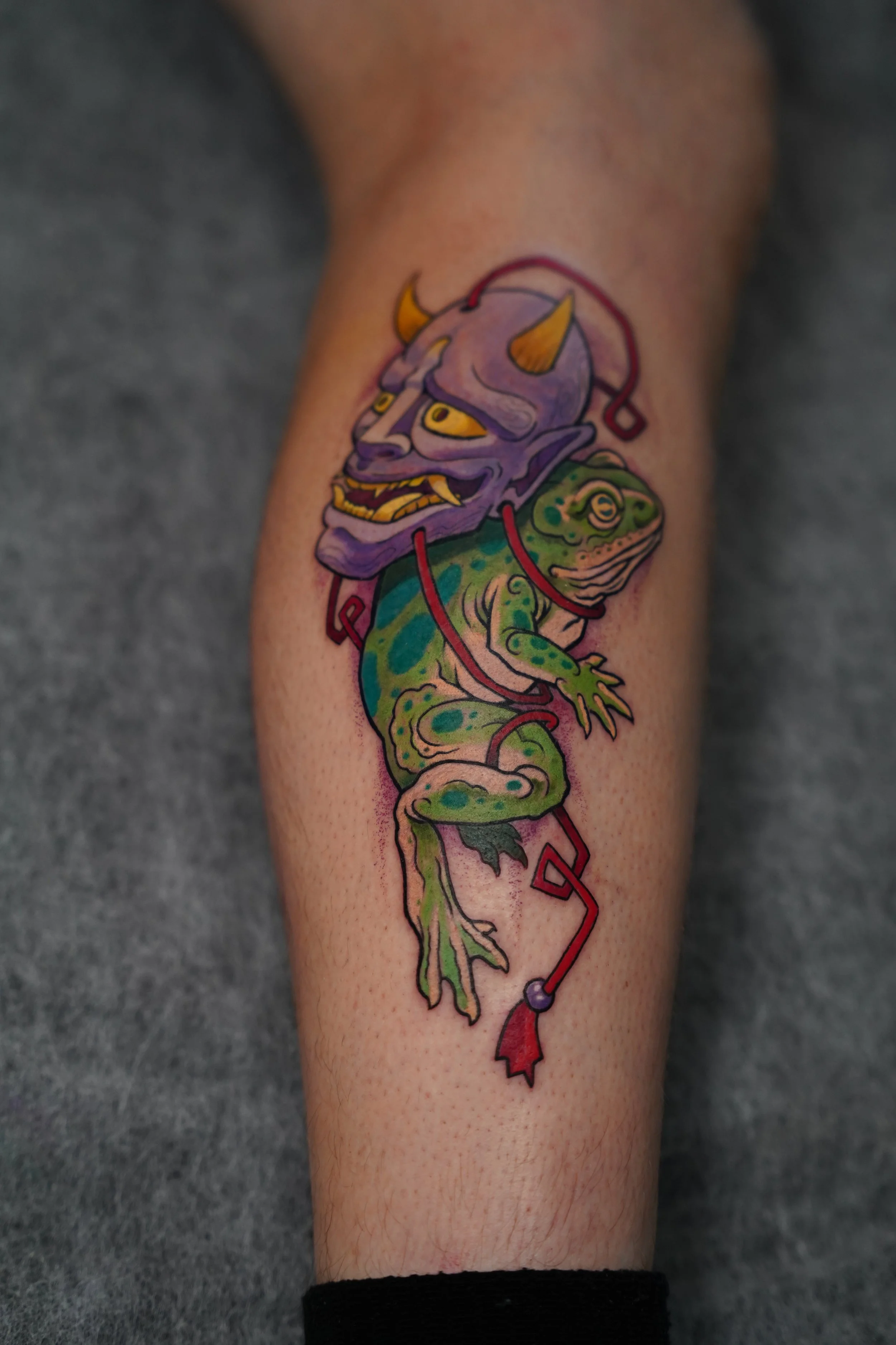 Tattoo of a creature with a purple demon-like face with horns, yellow eyes, and sharp teeth, attached to a green frog with blue spots, connected by red wires.