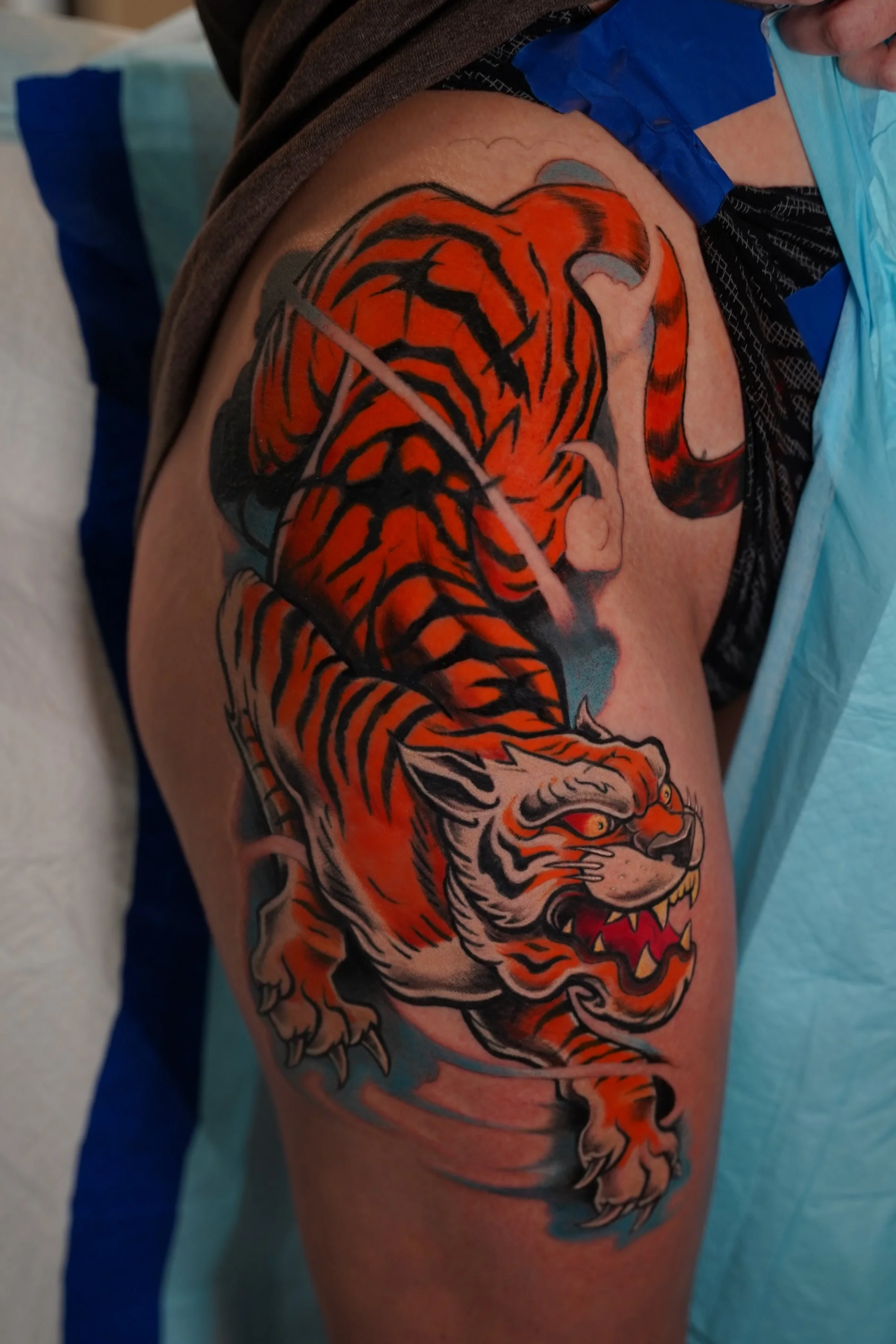 A colorful tattoo of a tiger on a person's upper arm and shoulder, featuring bold black and orange stripes, a fierce expression, and detailed claws.