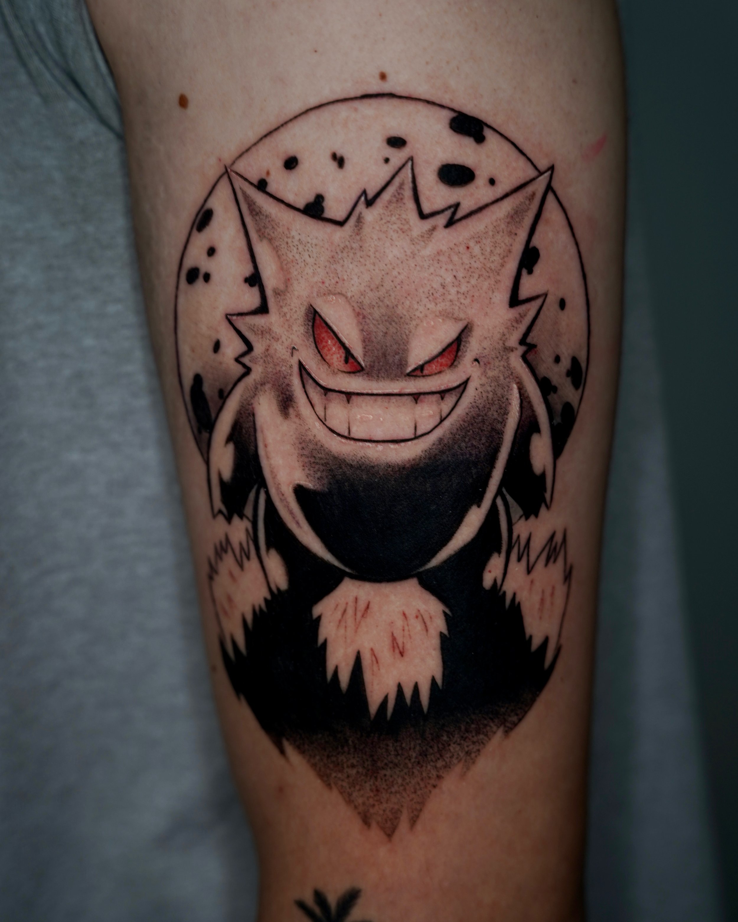 Tattoo of a mischievous, smiling creature with red eyes and sharp teeth, surrounded by a dotted circle, on a person's arm.
