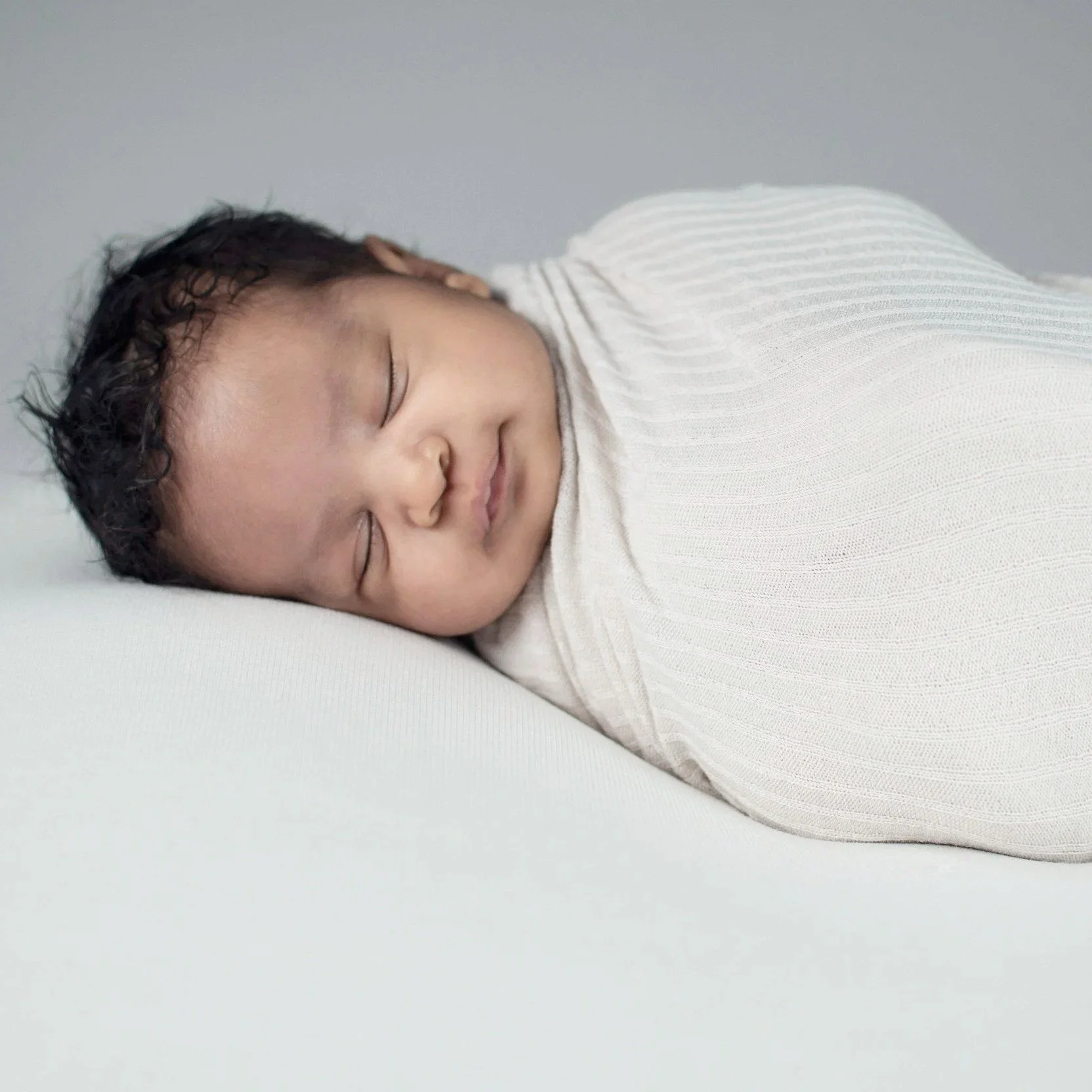 A sleeping baby with curly hair lying on a white surface, wearing a white outfit.
