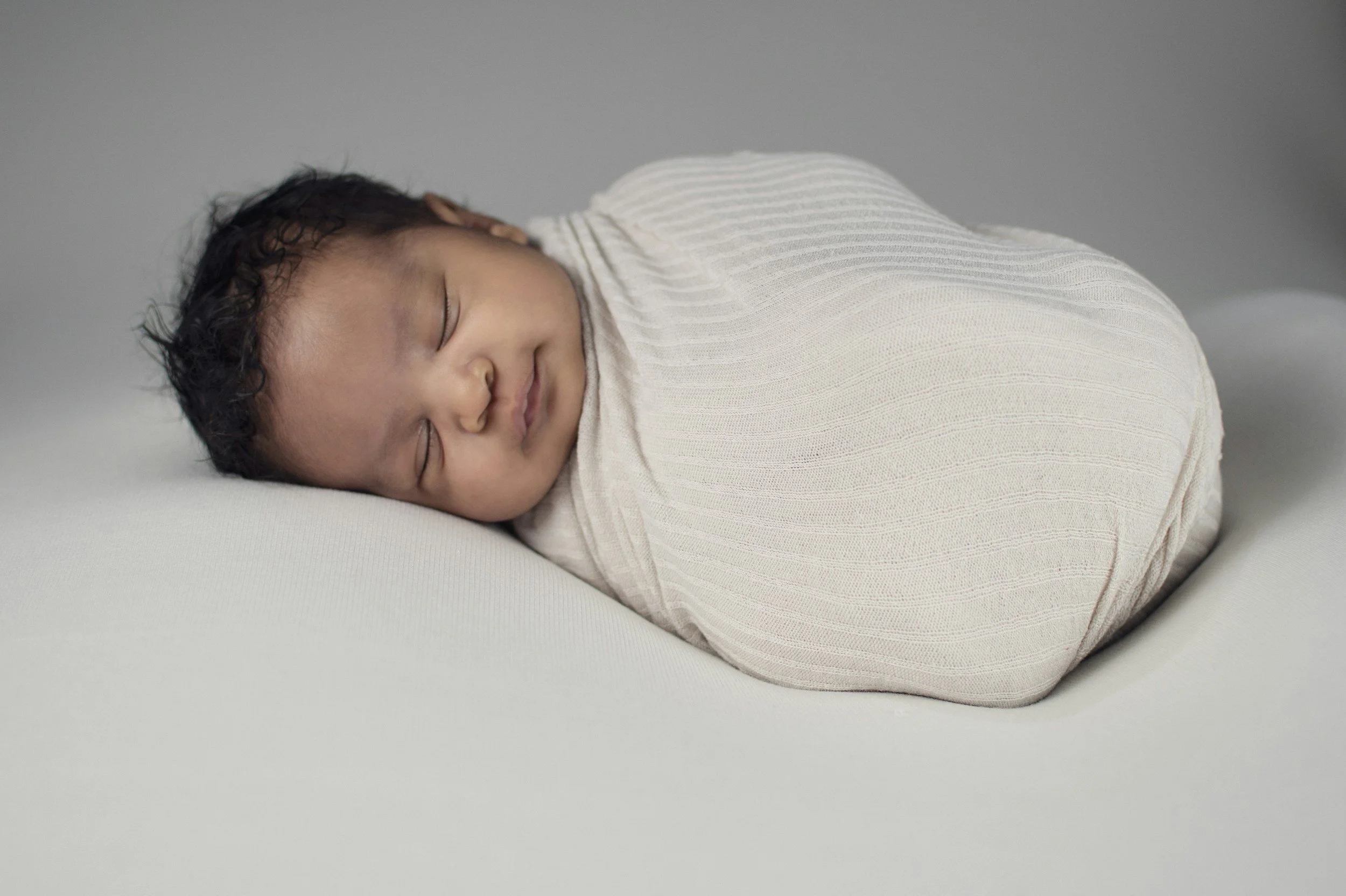 A sleeping baby wrapped in a white swaddle, lying on a white surface with a gray background.