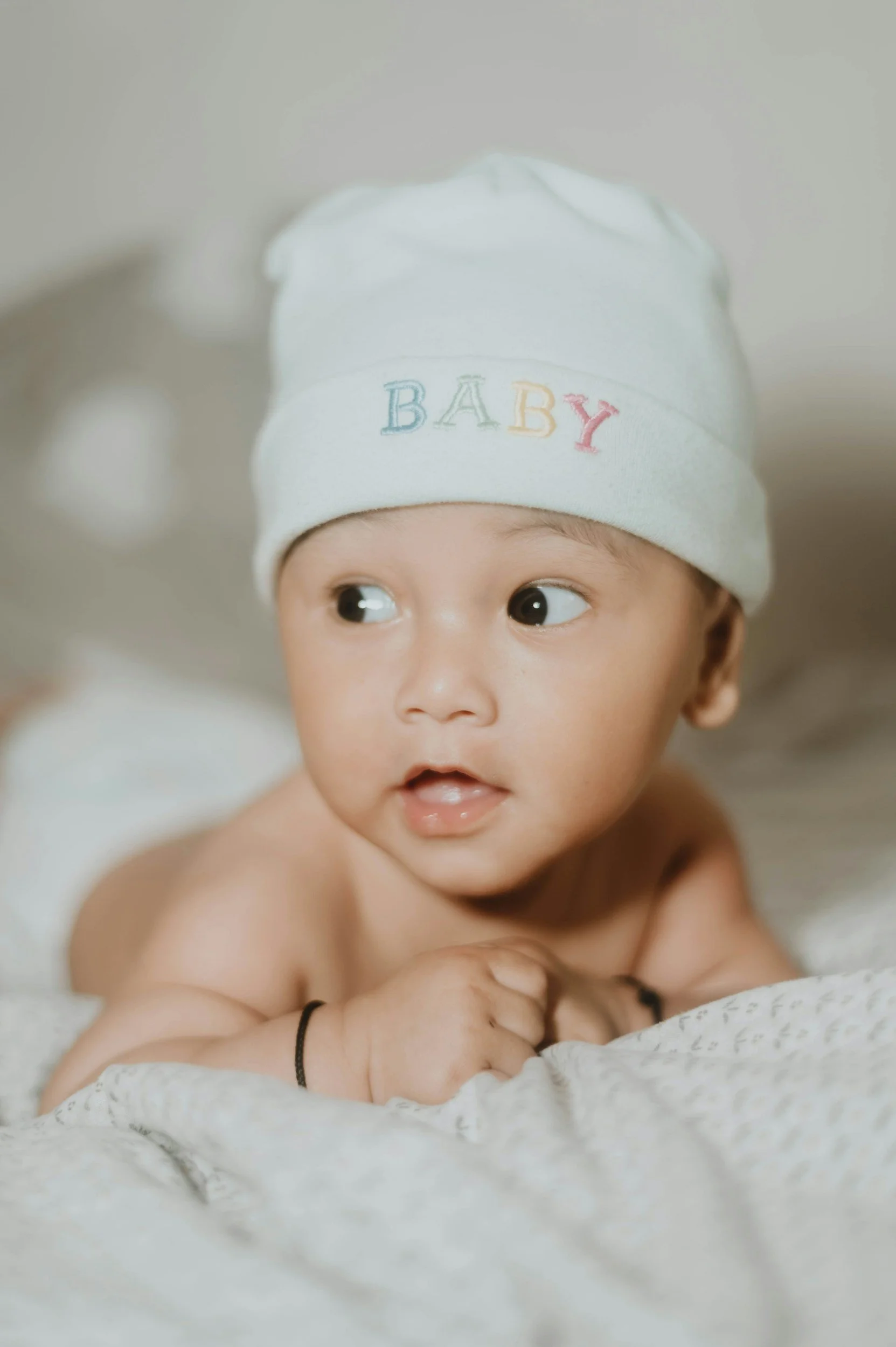 A baby lying on a bed, wearing a white hat with the word" BABY" embroidered in colorful letters.