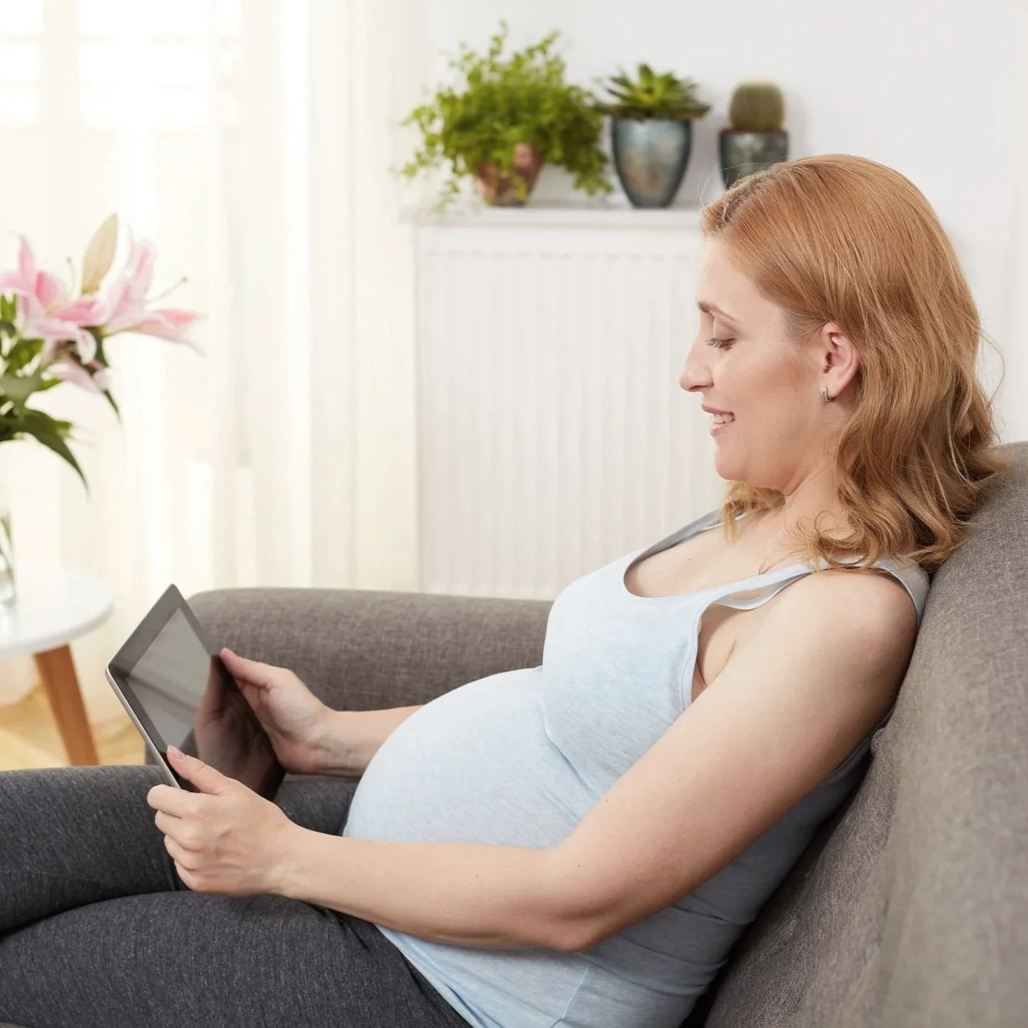 Pregnant woman with red hair smiling while sitting on a gray sofa, looking at a tablet she is holding.