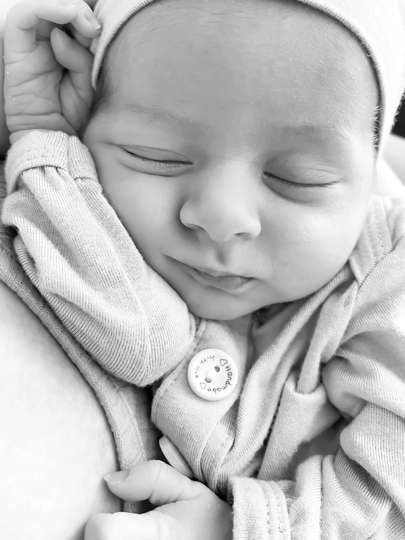 A close-up black and white photo of a sleeping baby with closed eyes, wearing a hat and a zip-up outfit, resting comfortably on someone's arm.