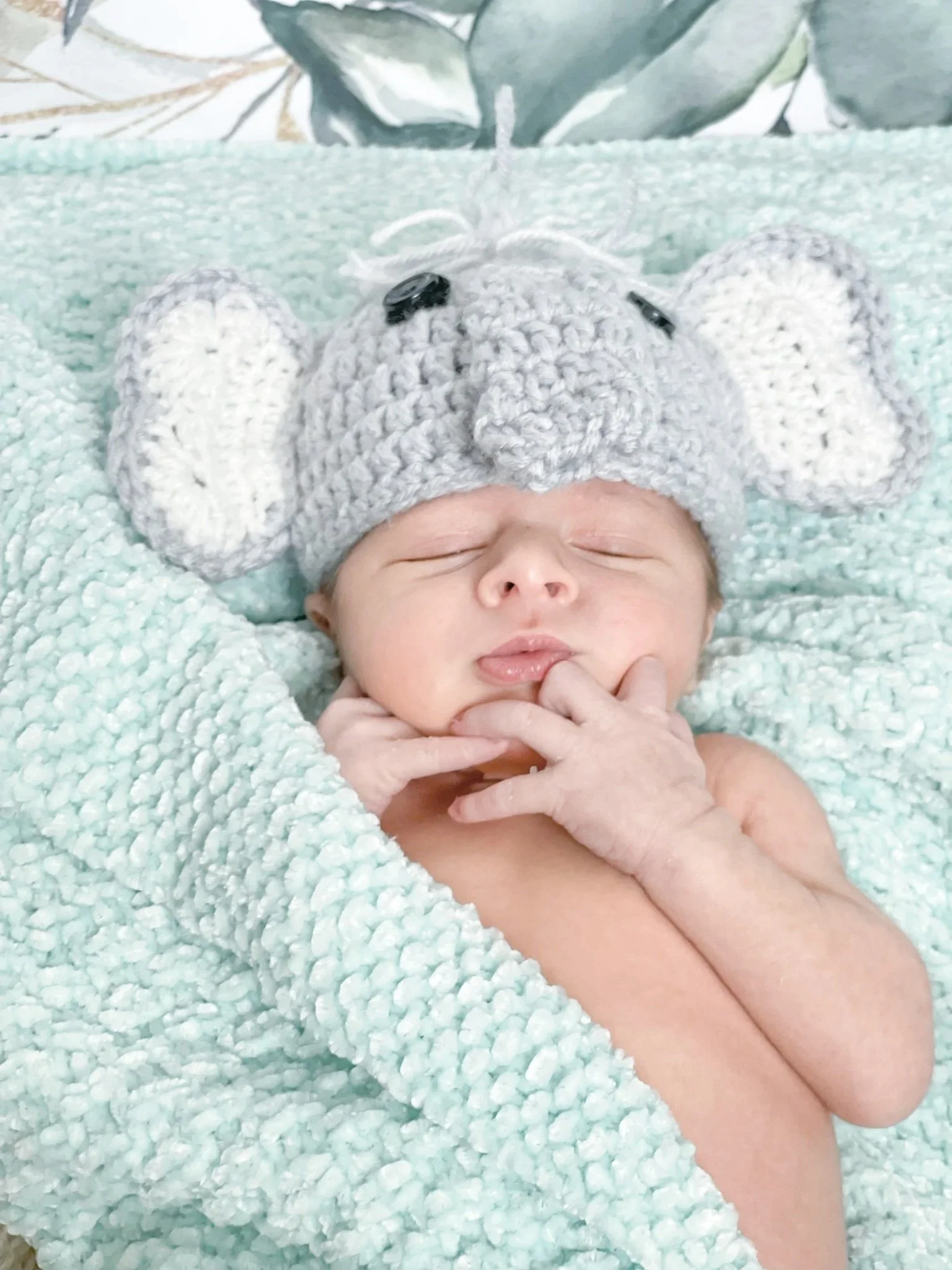 A newborn baby sleeping peacefully, wearing a gray crocheted mouse hat with floppy ears, surrounded by a soft mint-green blanket.