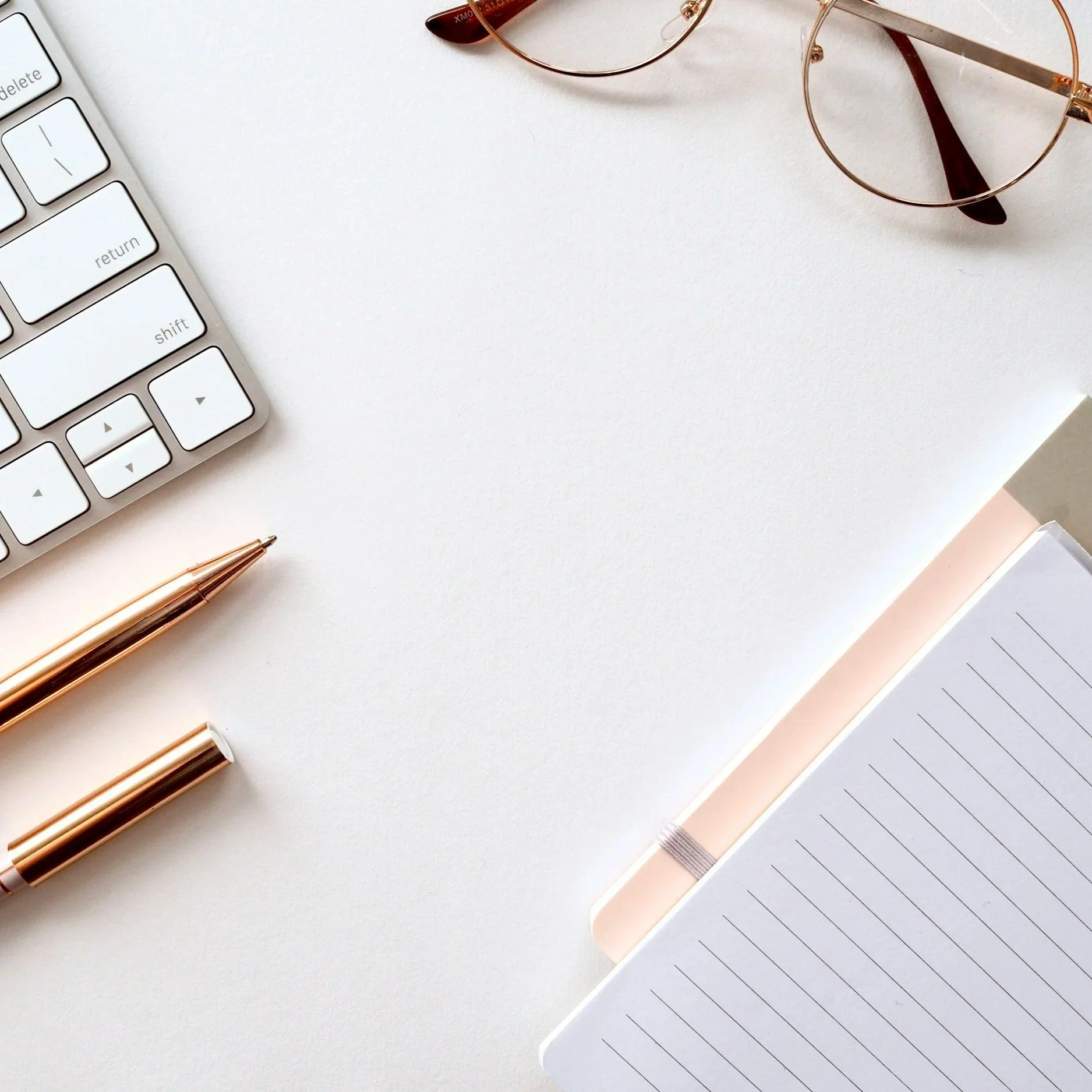 Flat lay of a white desk with a gold pen, a white keyboard, a pair of rose gold eyeglasses, a notebook with a pink cover, and a lined notepad.