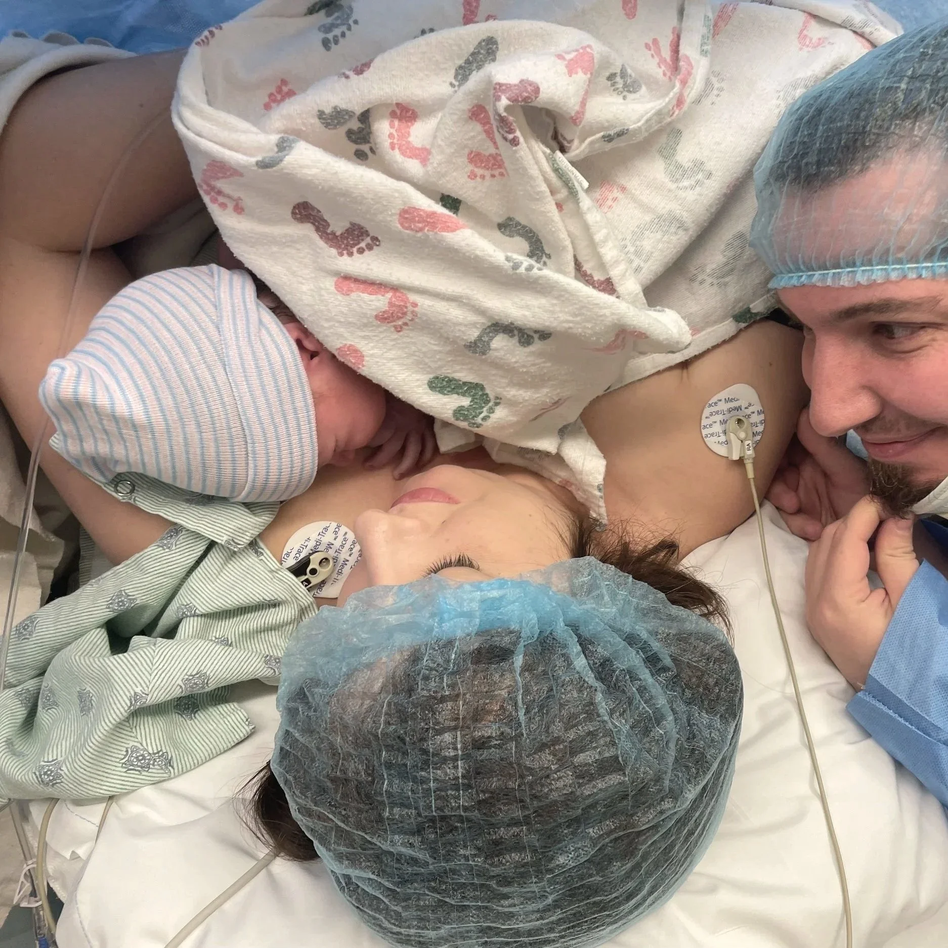 A mother lying in a hospital bed holding her newborn baby close. Both are wearing hospital caps, and the mother has electronic monitoring pads on her chest and arm. The scene depicts a moment of bonding shortly after childbirth.