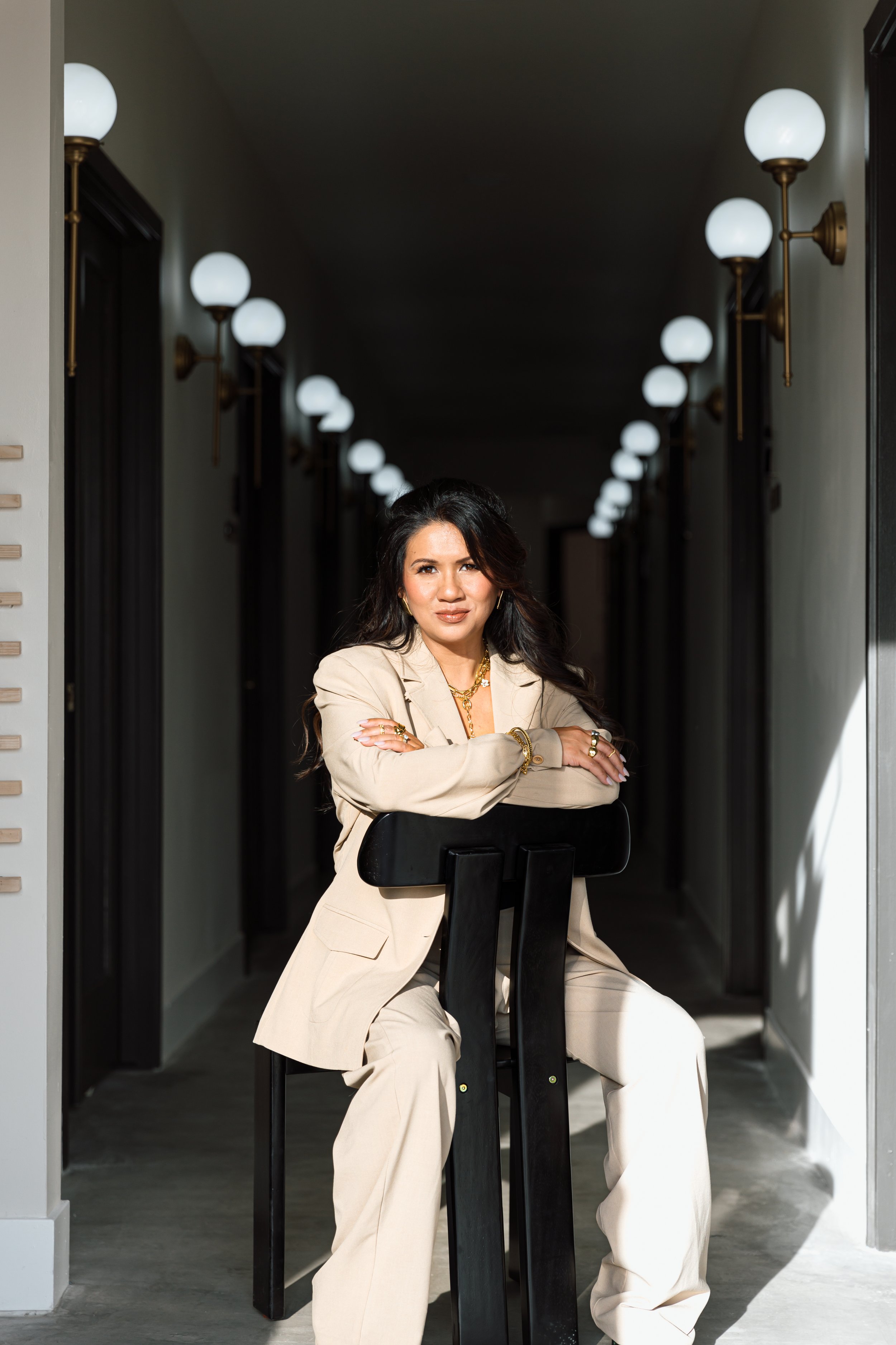 A woman dressed in a beige pantsuit and jewelry sitting on a black stool in a corridor with black walls and vintage wall sconces with white globes.