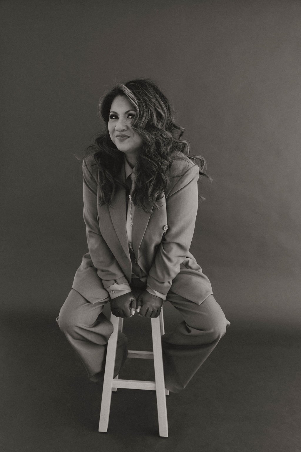 Black and white photo of a woman with long, wavy hair sitting on a stool, wearing a blazer, gloves, and pants, against a plain background.
