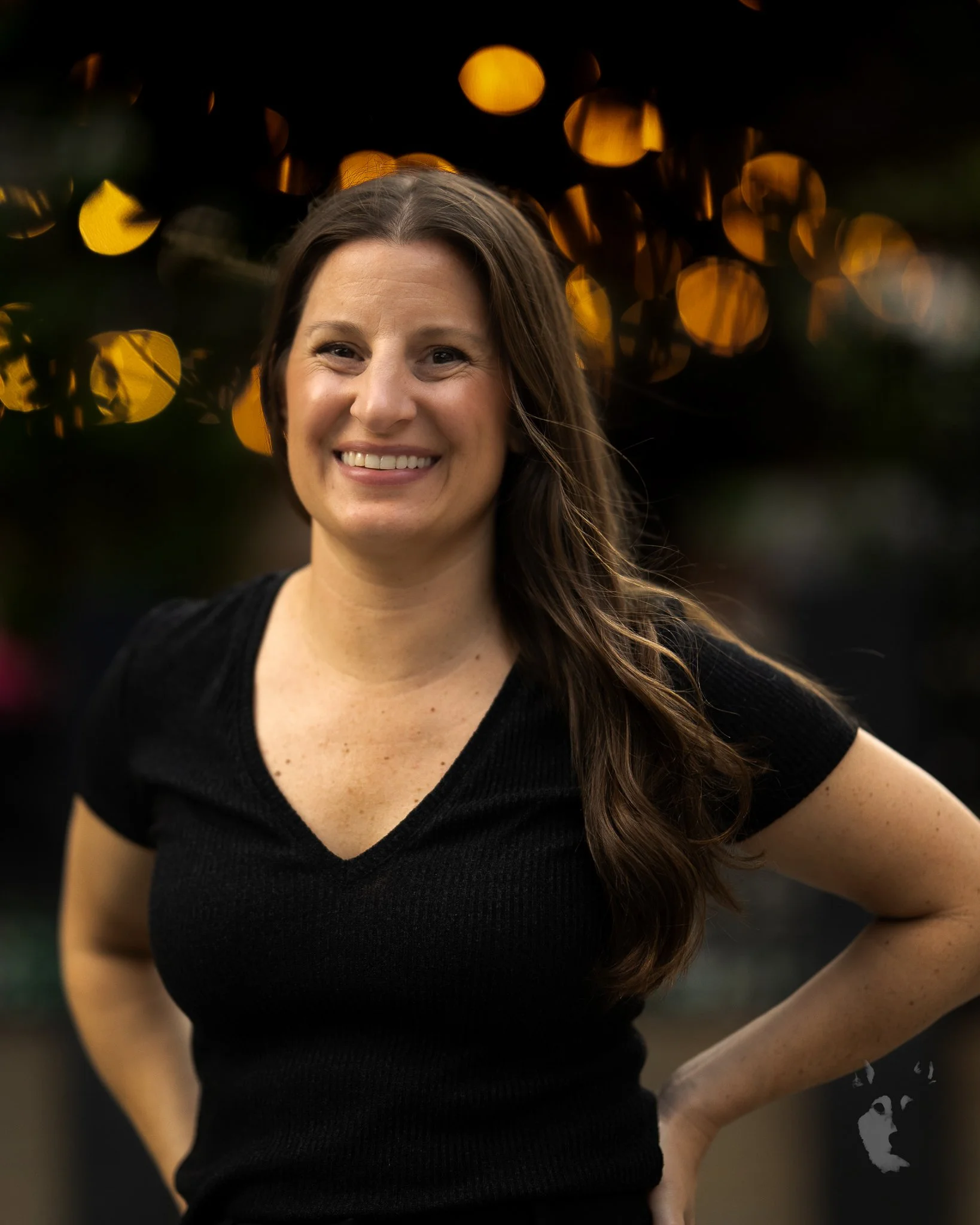 A smiling woman with long brown hair wearing a black V-neck shirt, standing outdoors at dusk with blurred yellow lights in the background.