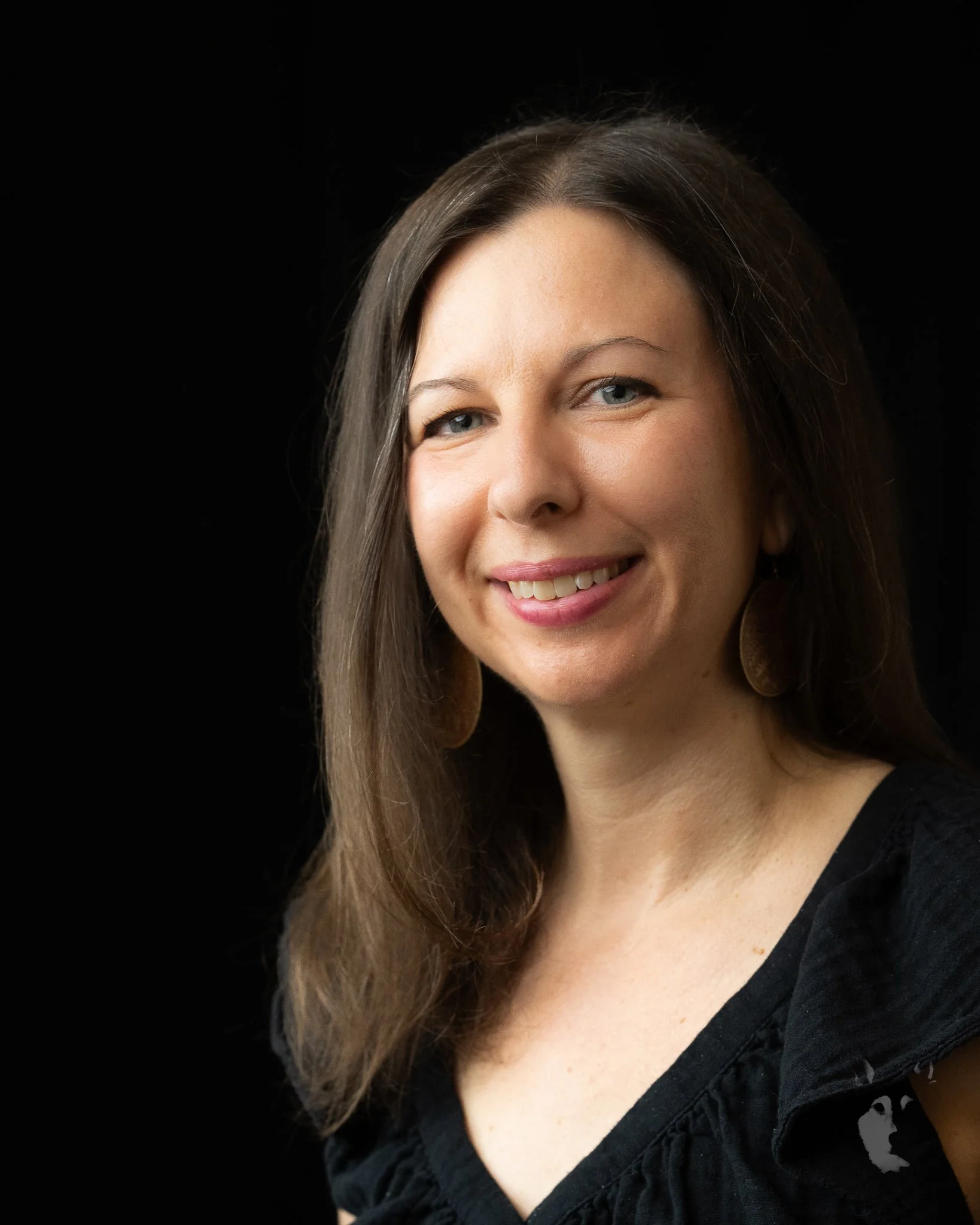 A smiling woman with long brown hair, wearing a black top and earrings, against a black background.