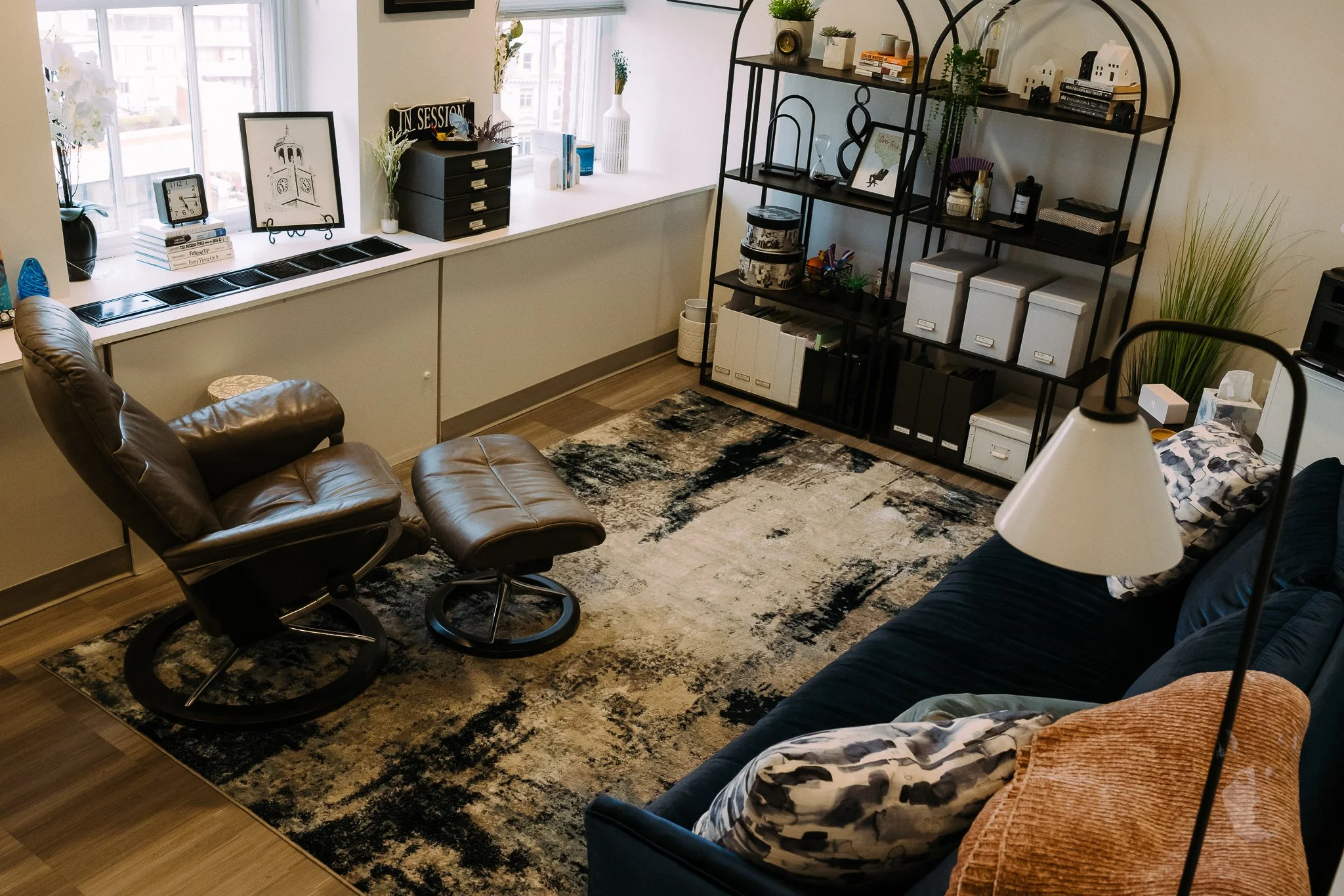 A cozy living room with a leather rocking chair and matching ottoman near a window with a white sill. The sill has books, a round clock, and decorative items. There's a dark blue sofa with pillows and a tall floor lamp nearby. Against the wall, a black metal shelving unit holds storage boxes, decorative objects, books, and plants. A multicolored abstract rug covers the wooden floor.