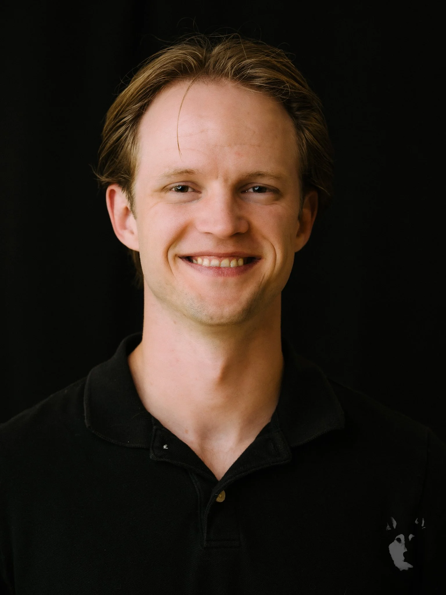 A smiling young man with light brown hair, wearing a black collared shirt, standing against a black background.