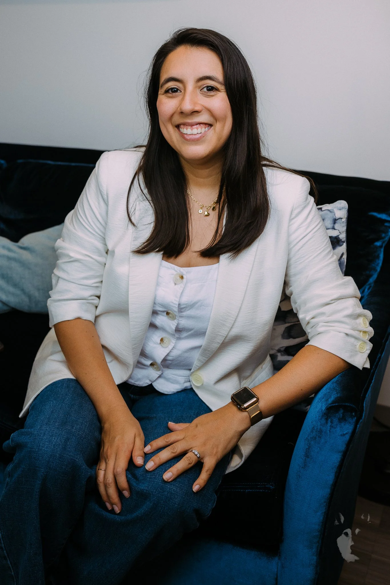 A woman with long dark hair smiling and sitting on a black velvet couch, wearing a white blazer, white top, blue jeans, a watch, a ring, and a necklace.