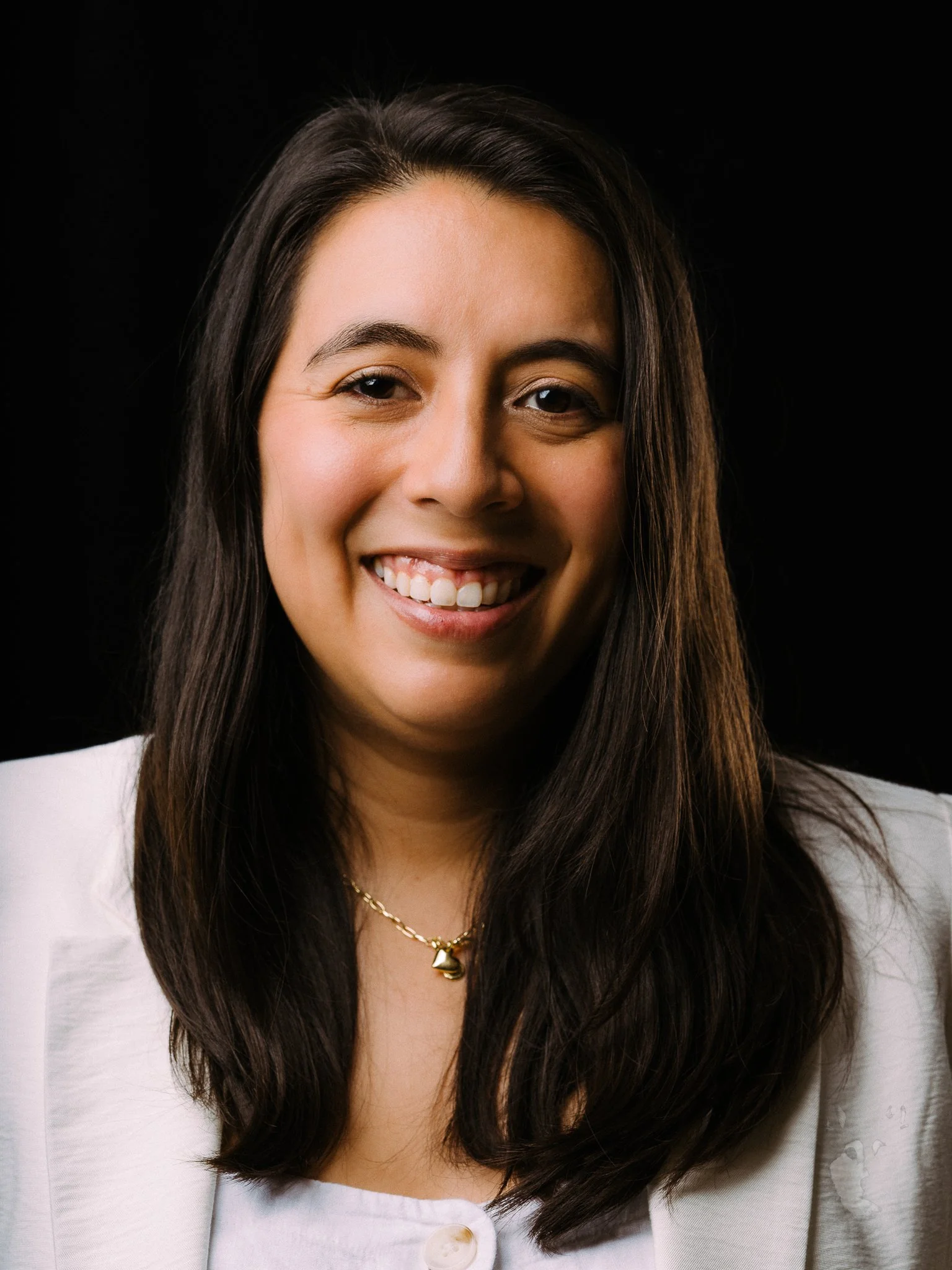 Close-up portrait of a smiling woman with dark brown hair, wearing a white blazer and gold necklace, against a black background.