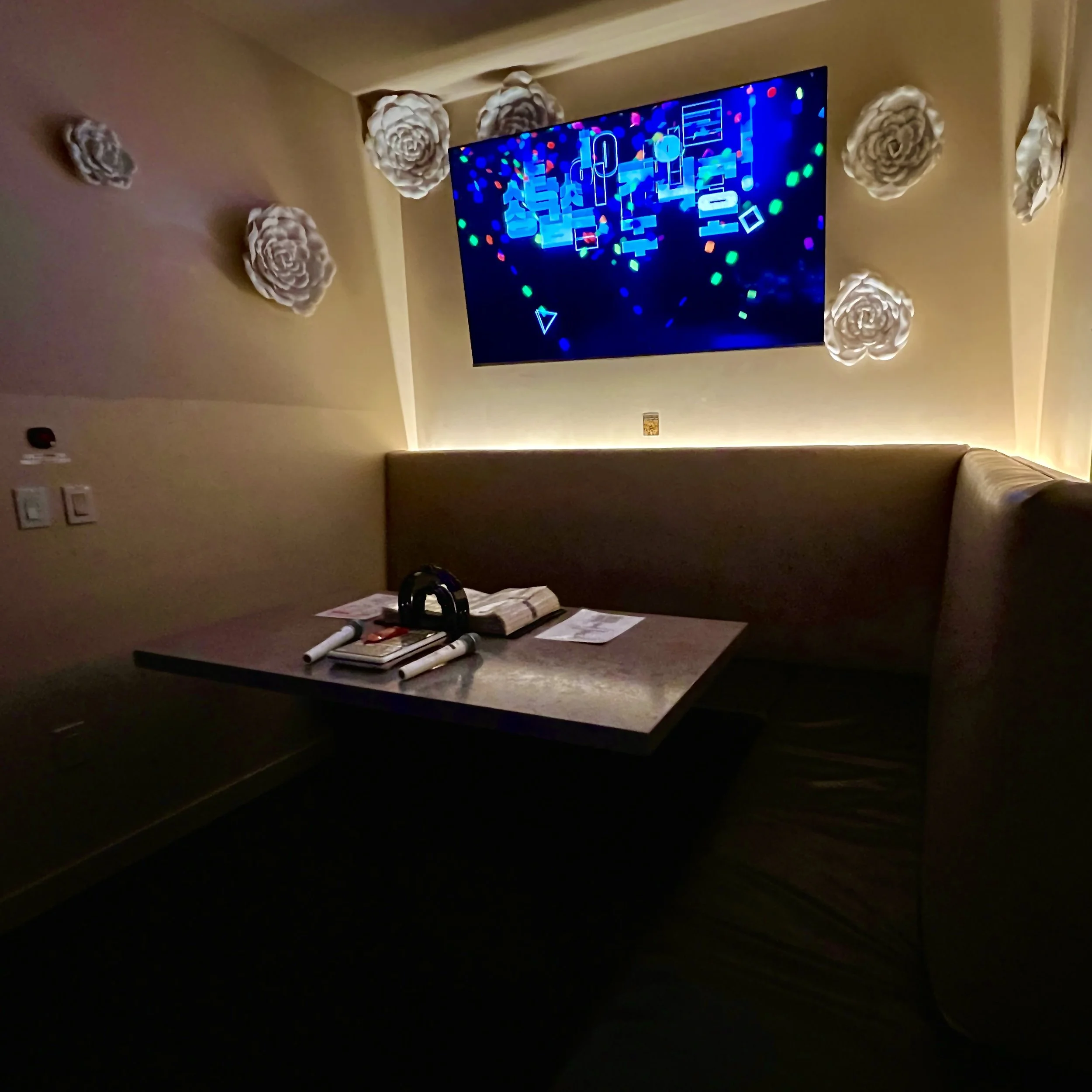 A dimly lit karaoke booth with a television screen displaying colorful graphics. There are decorative white paper flowers on the beige walls. A table in the booth has a black microphone, papers, and a remote control.