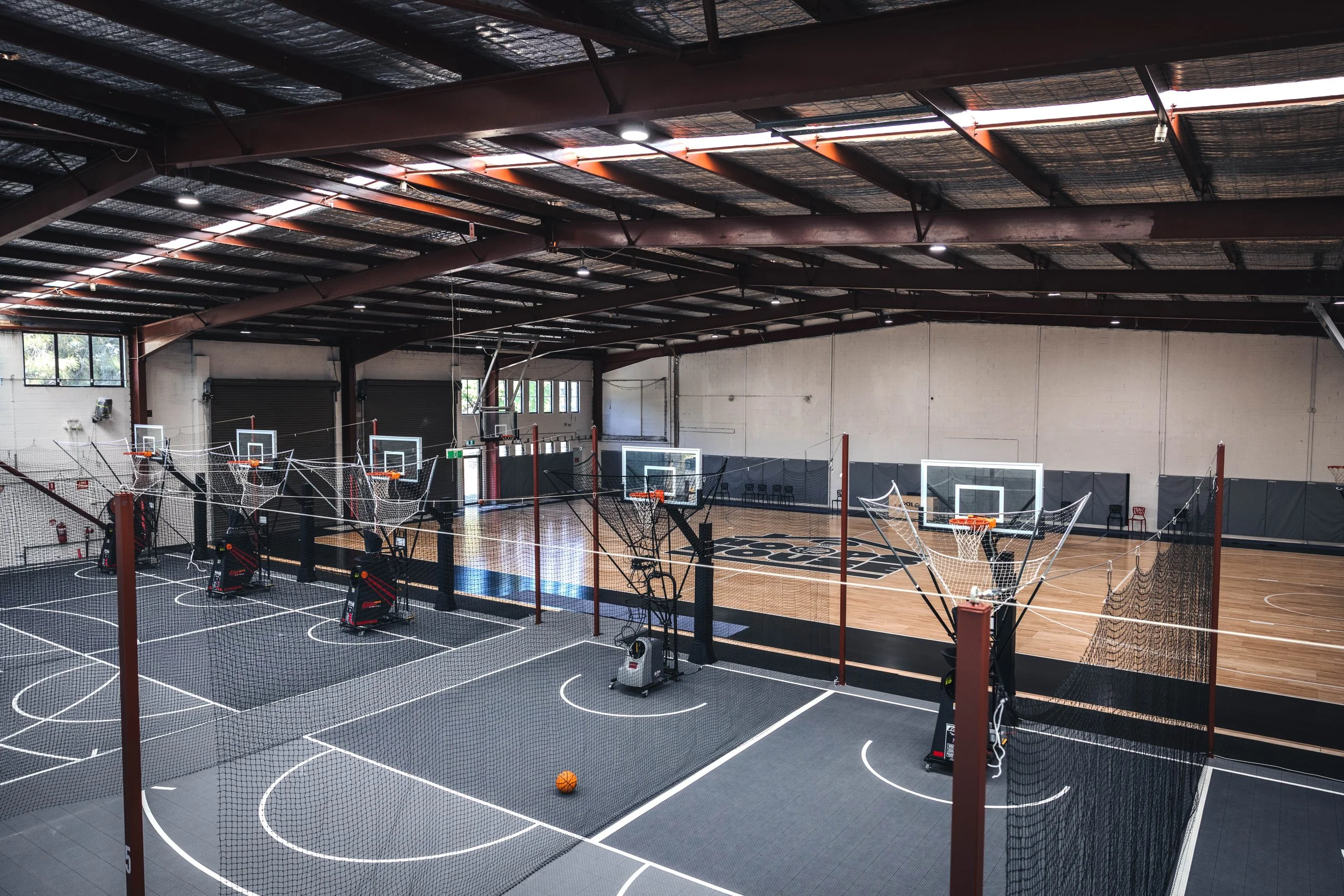 Indoor basketball court with multiple hoops and a small basketball on the court. The court has a black border and the basketball area is visible. The ceiling has industrial-style beams and skylights, and there are fencing and padding along the walls.