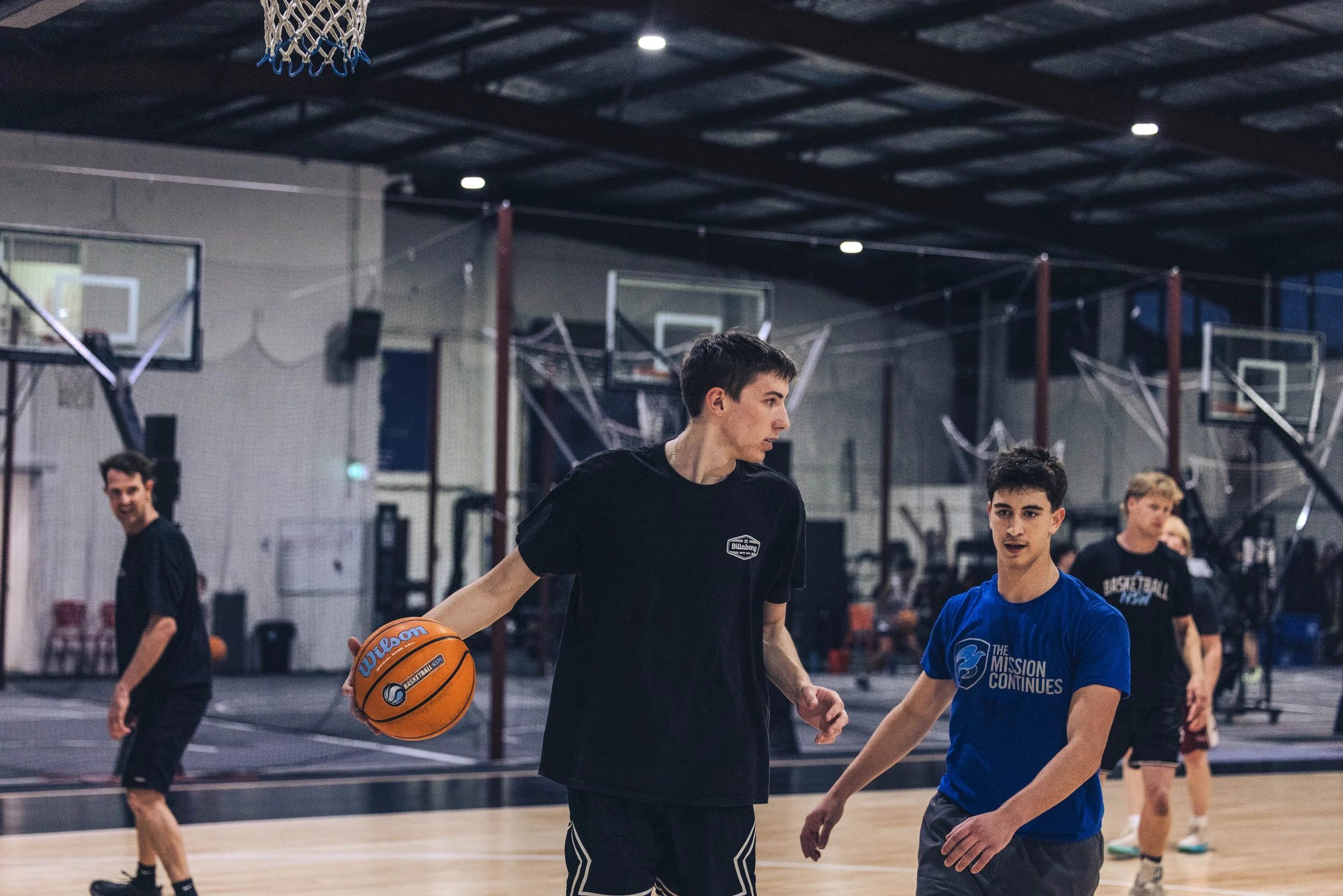 Indoor basketball court with several players, including one holding a basketball, engaged in a practice or game, with basketball hoops and court markings visible.