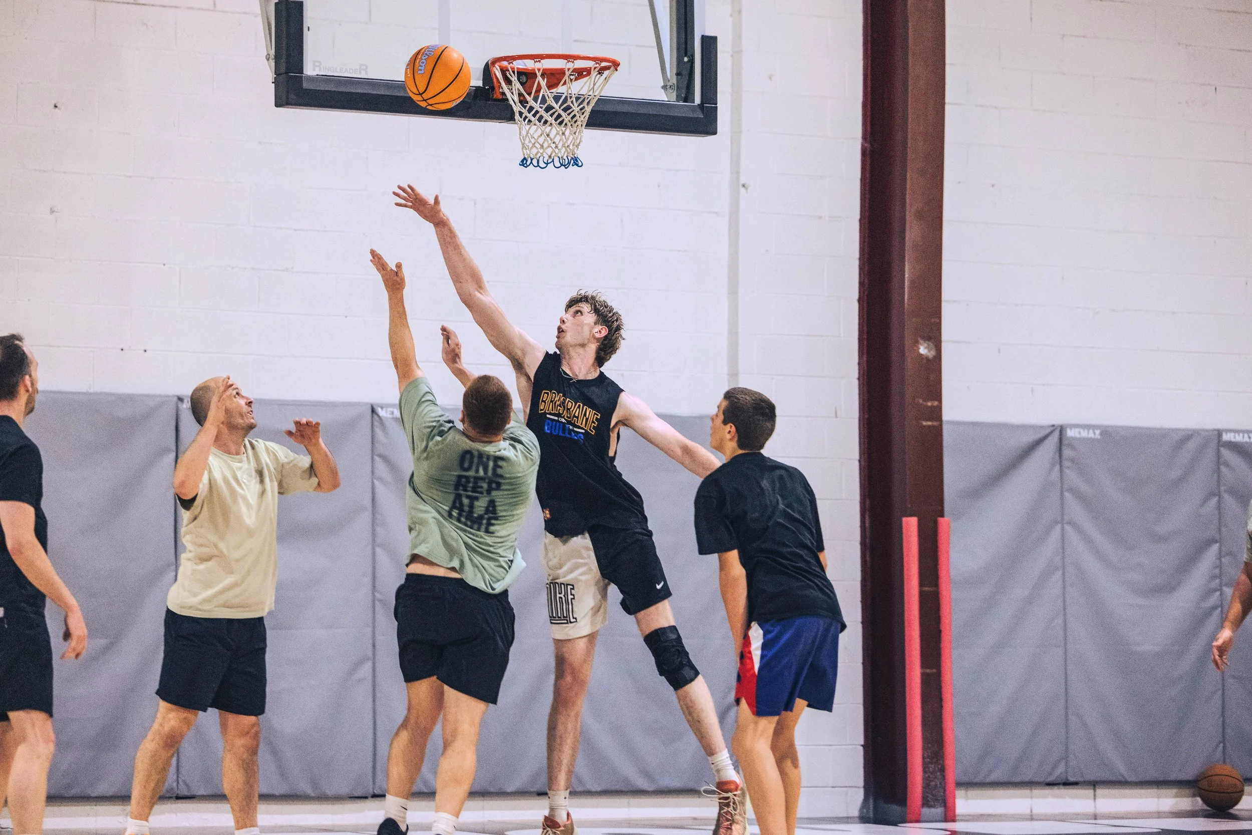 A group of people playing basketball in a gymnasium, with one player attempting a shot near the hoop while others try to block or defend.