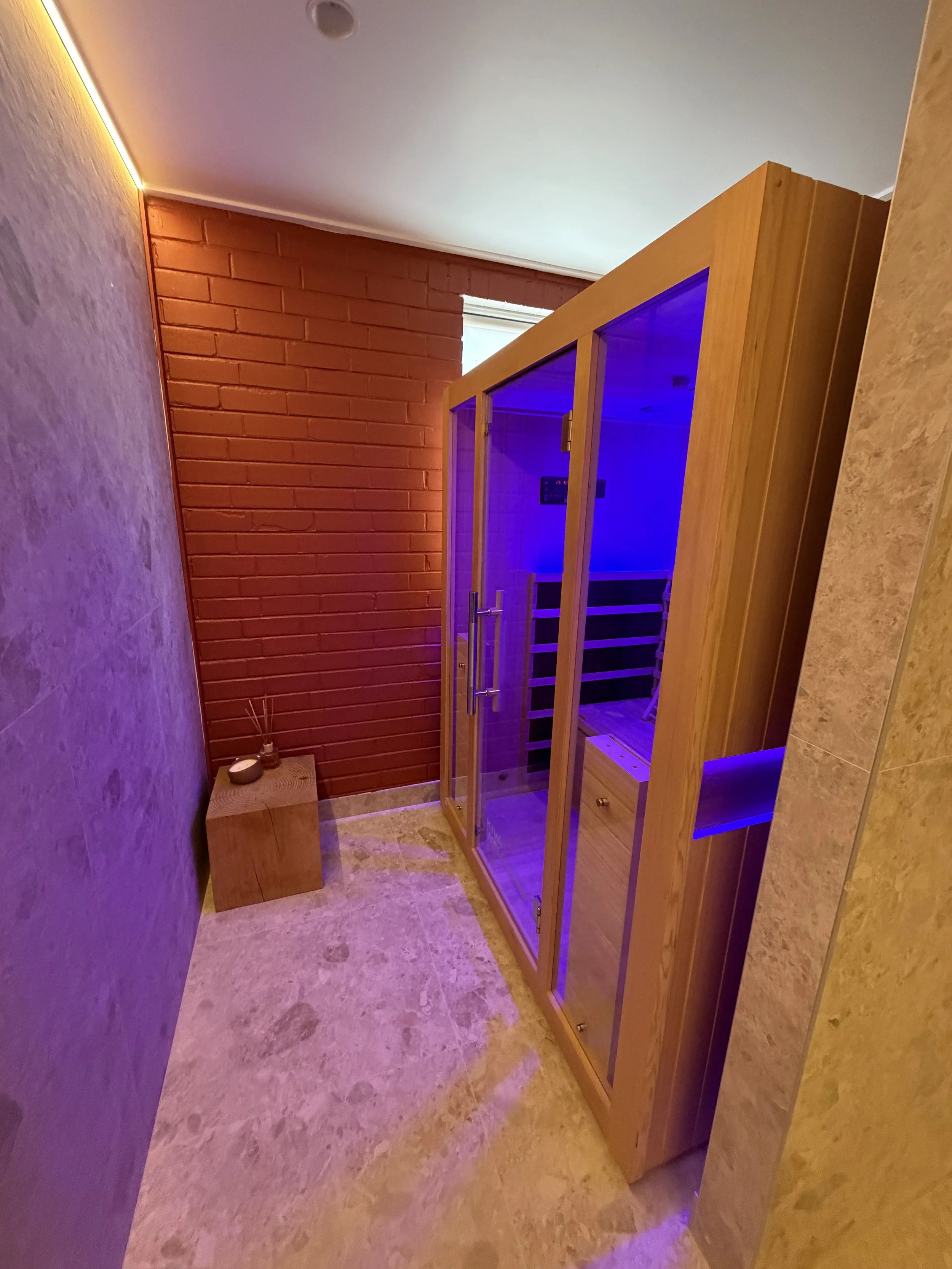 A small room with a wooden infrared sauna in the corner, purple lighting inside, a textured brick wall, and a small wooden side table with a diffuser.