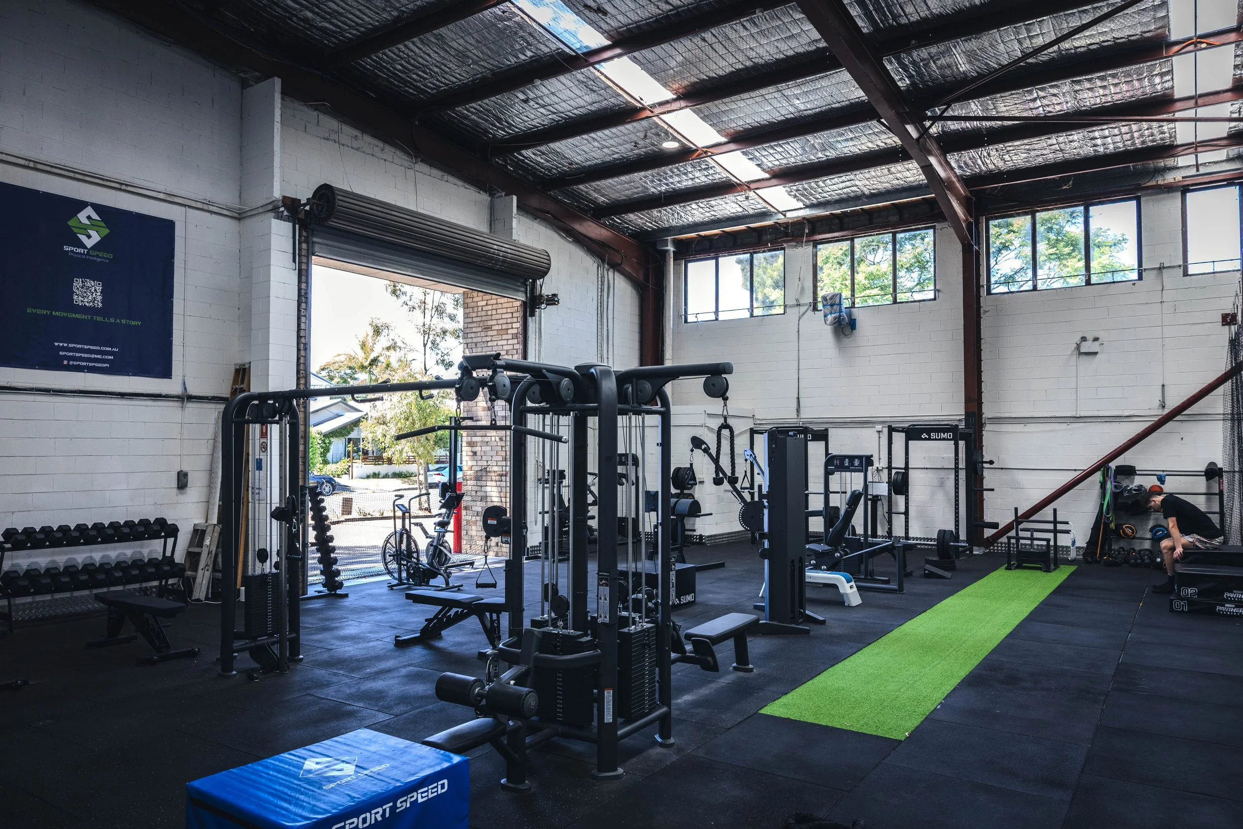 Interior view of a gym with various exercise equipment, including weights, machines, and a green turf strip, with a high ceiling and large windows letting in natural light.
