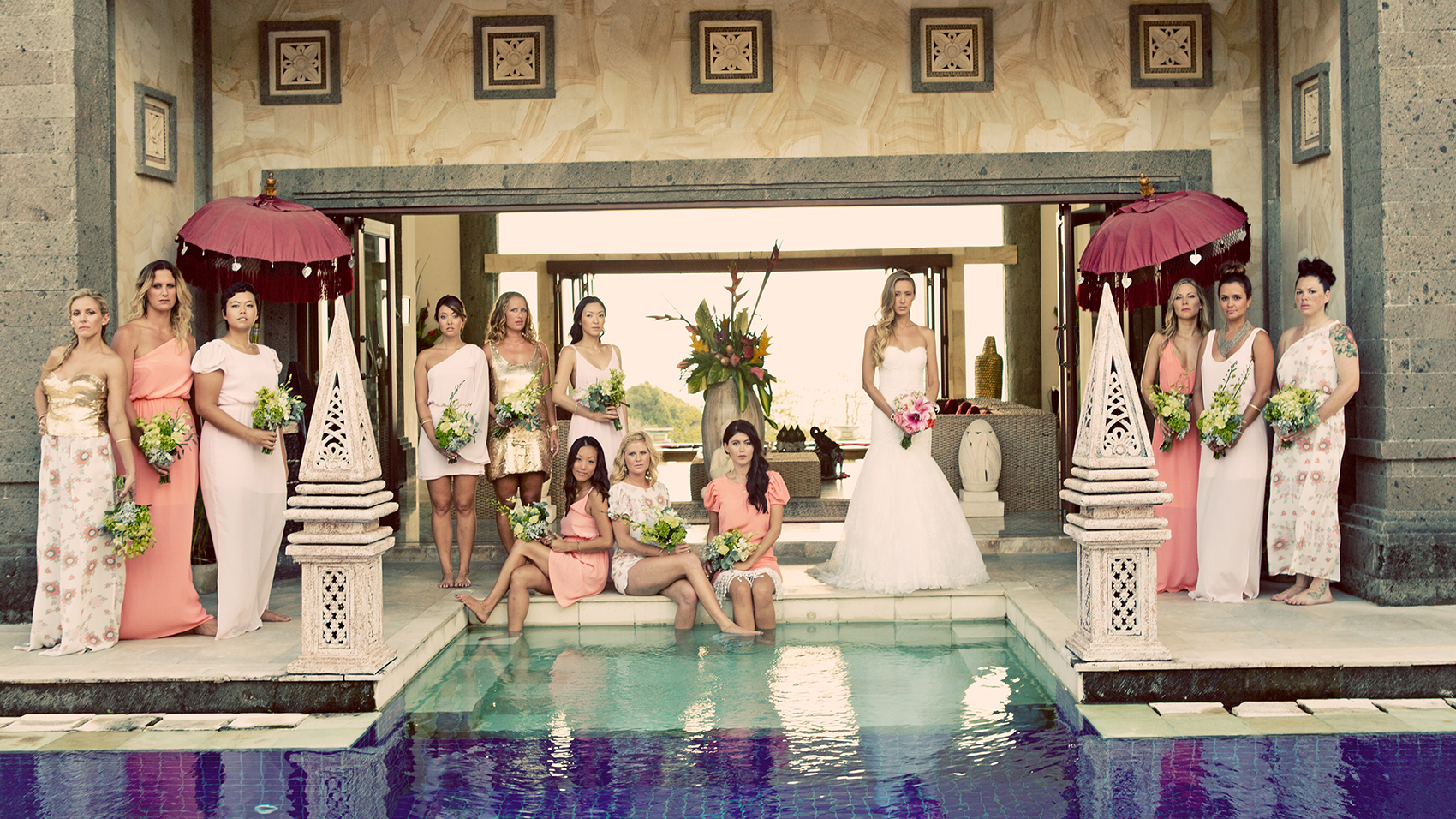 A group of women in formal dresses, some holding flowers, standing around a swimming pool with a woman in a white wedding dress holding pink flowers at the center, under two large red umbrellas, inside a decorative indoor setting.
