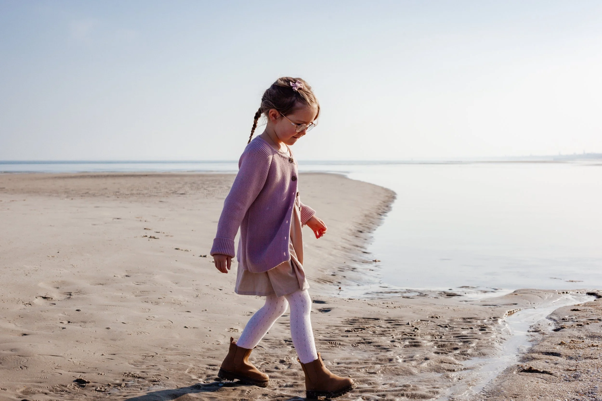 A young girl with glasses and braided hair walking along the sandy shoreline of a beach near the water.
