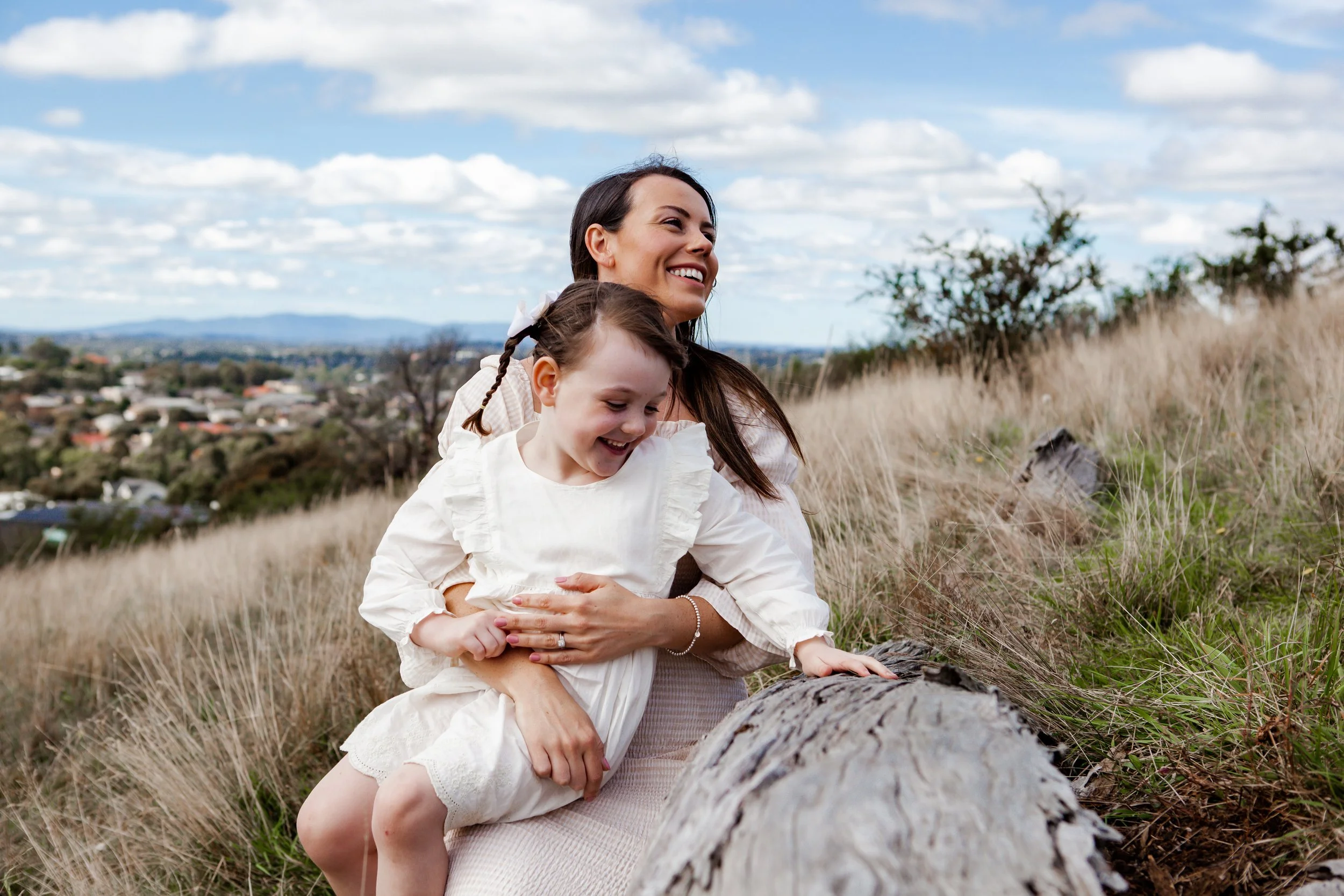 A woman and a young girl are outdoors on a grassy hillside, smiling and playing on a fallen tree trunk.