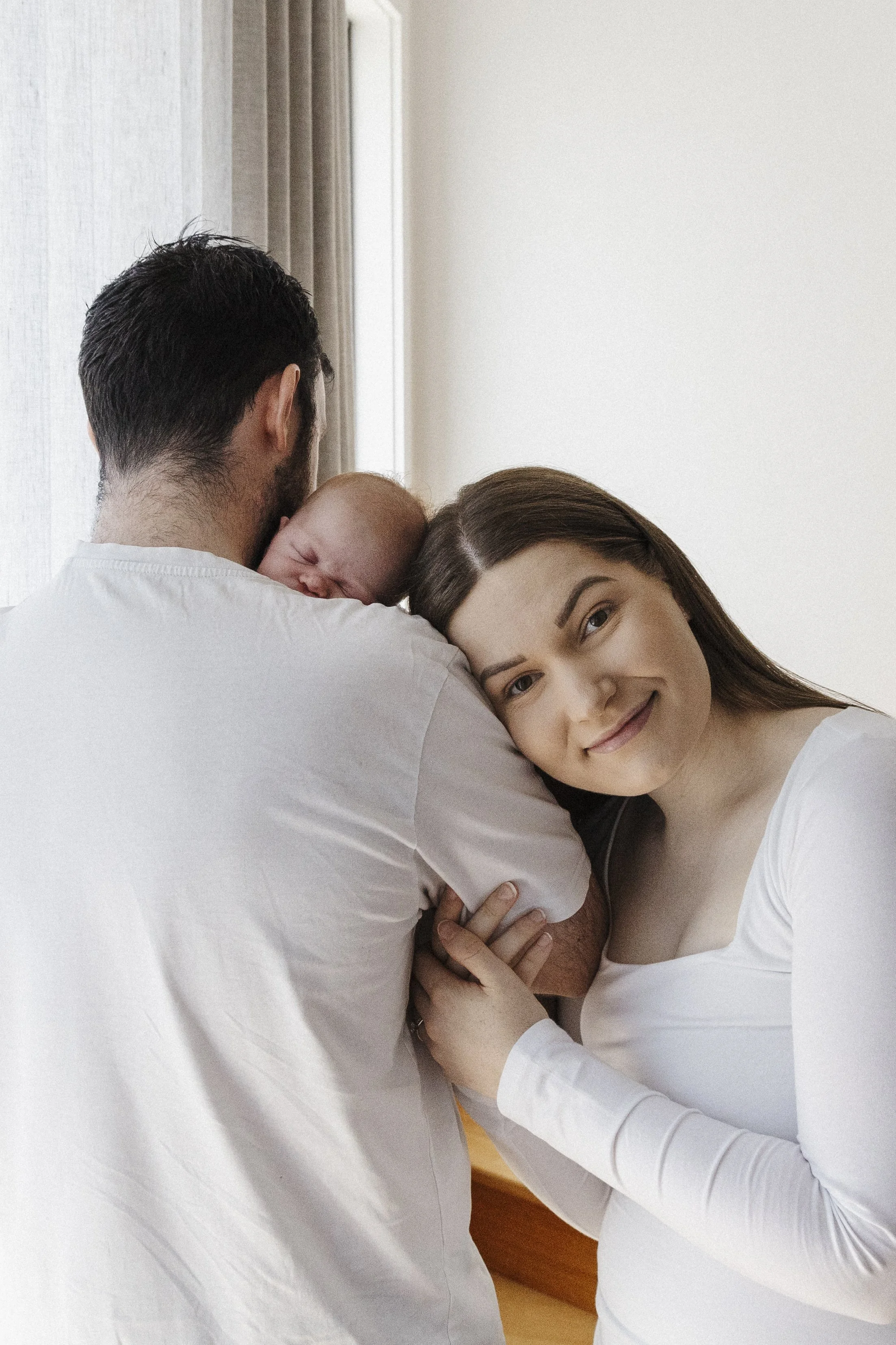 A woman is smiling while holding her newborn baby, who is resting on her husband's shoulder. The husband, with his back to the camera, has the baby on his shoulder, in a light-colored room with natural light.
