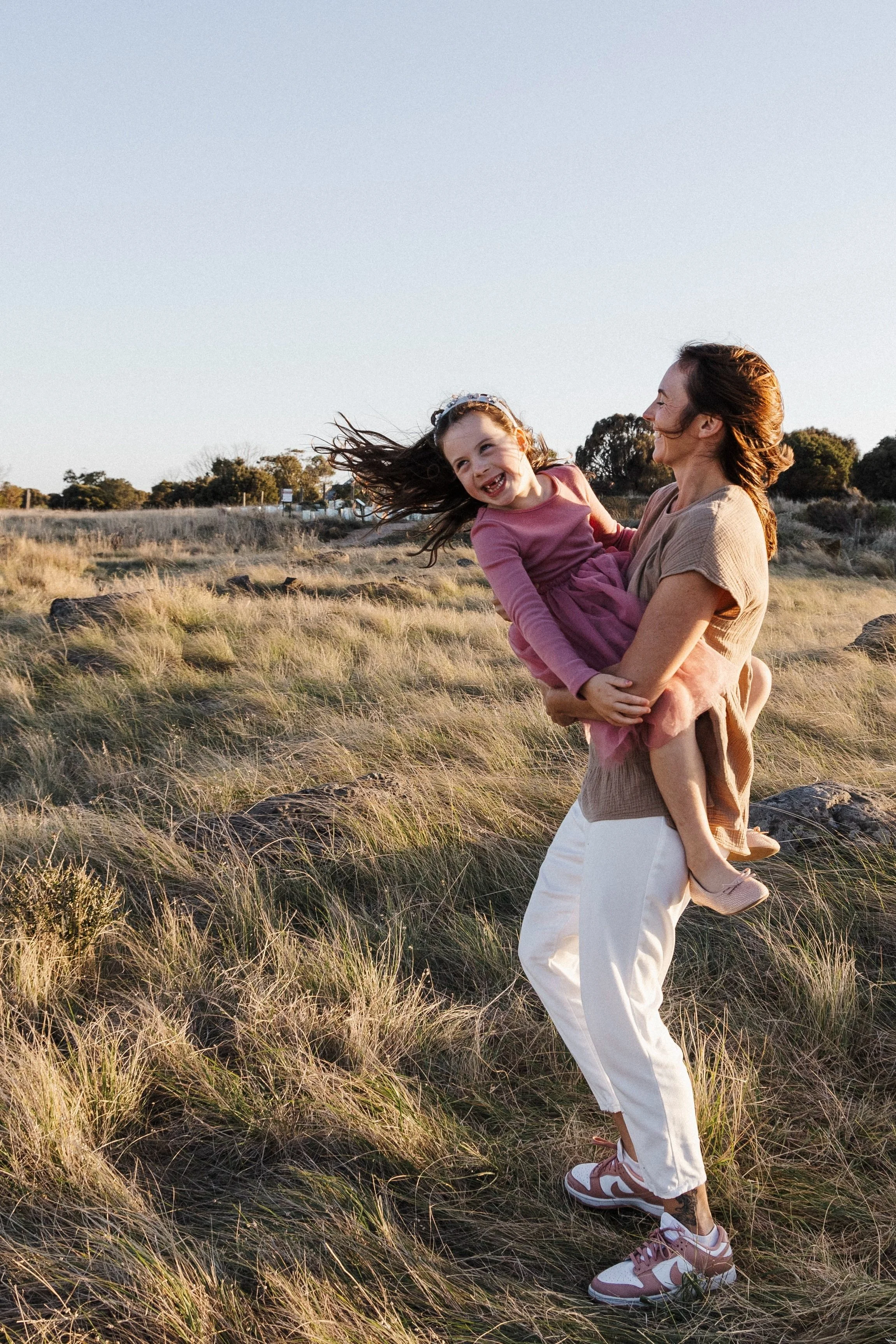 A woman in casual clothes lifts a young girl in a pink dress in a grassy field during the daytime. The girl is smiling and laughing, with her hair flowing in the wind. The background features trees and a clear sky.