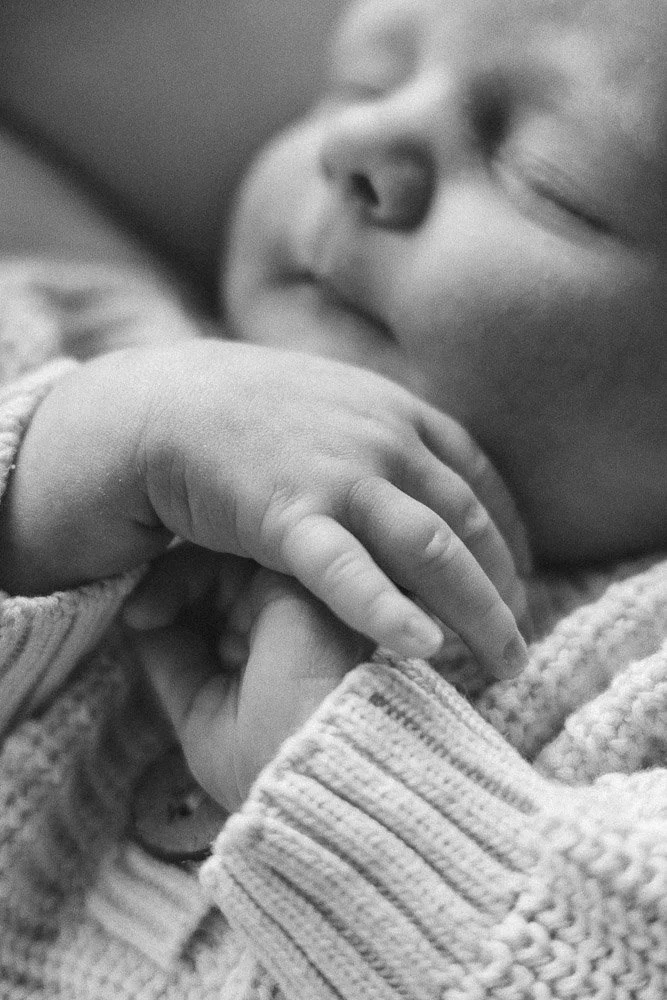Close-up of a sleeping baby holding an adult's finger, wearing a knitted sweater.