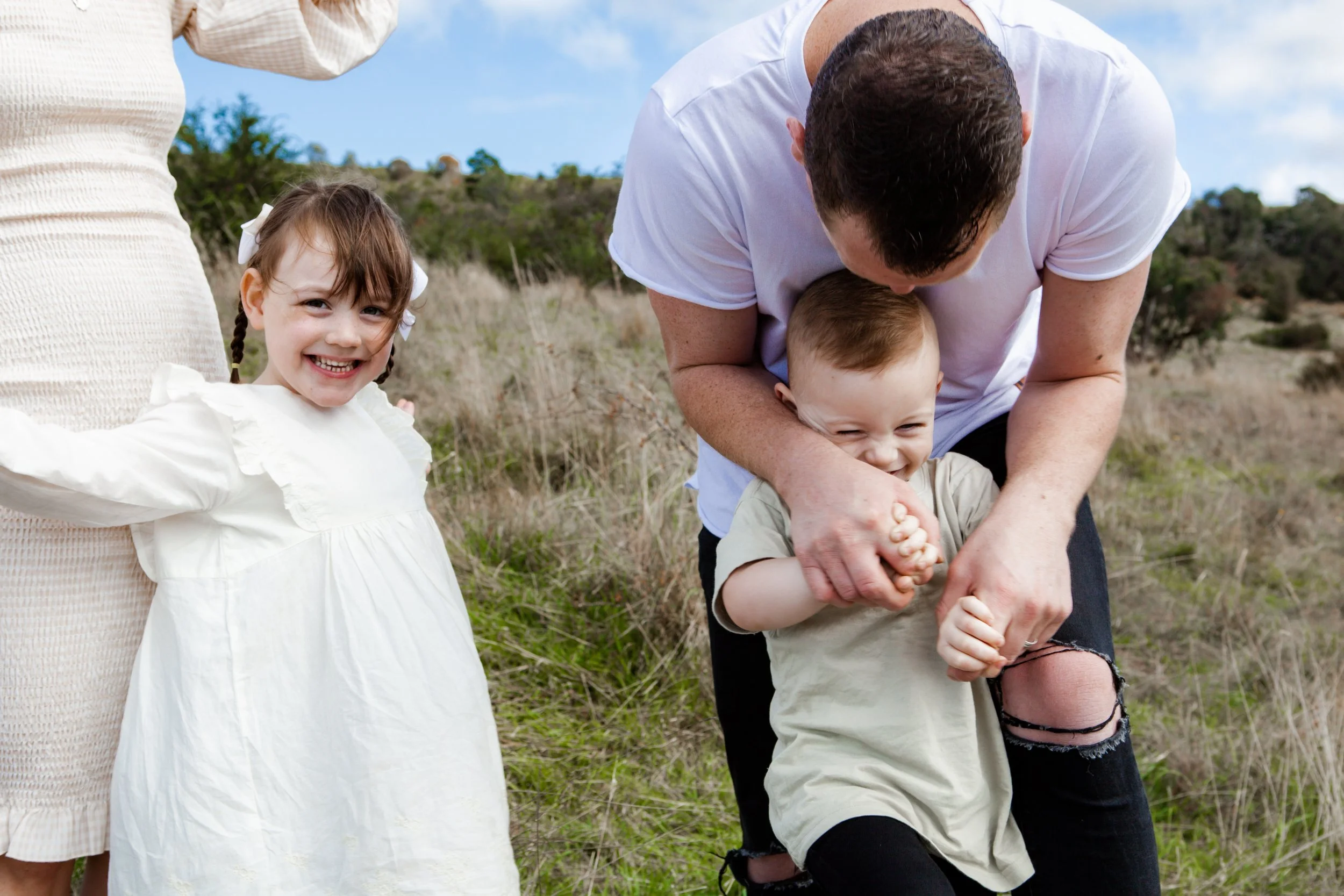A man is playfully wrestling a young boy while a girl in a white dress smiles nearby at the park.