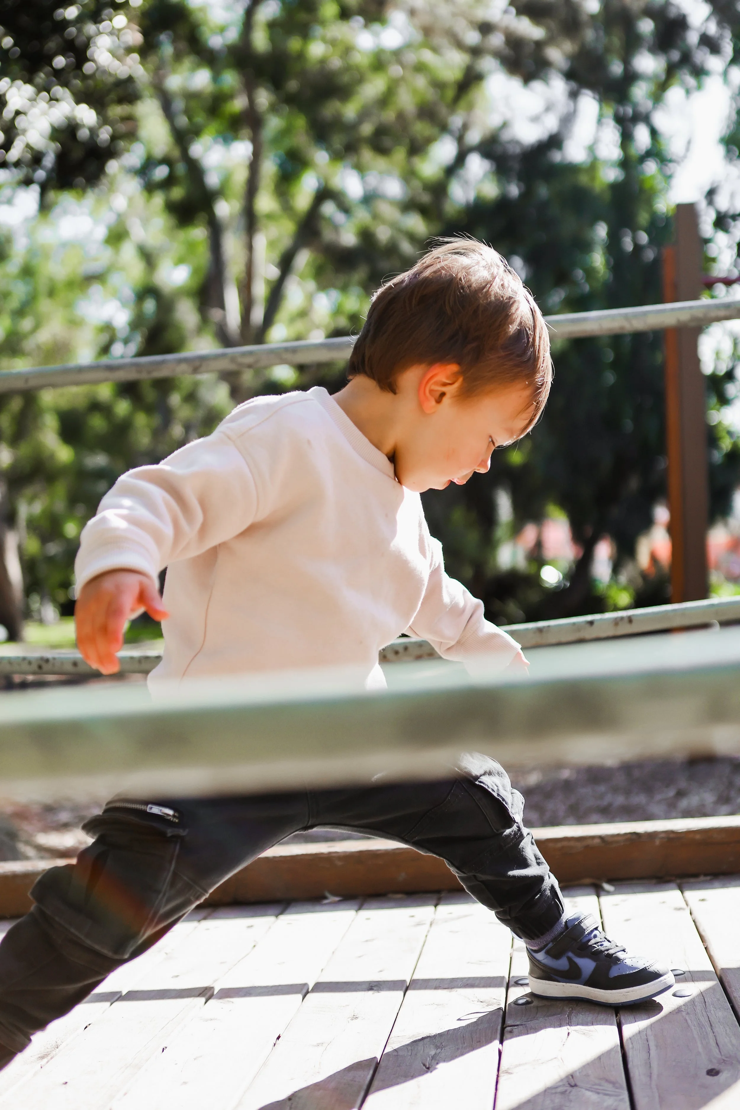 Young boy playing outdoors on a sunny day, leaning forward and reaching with his left hand, wearing a white sweatshirt, dark pants, and athletic shoes, with trees in the background.