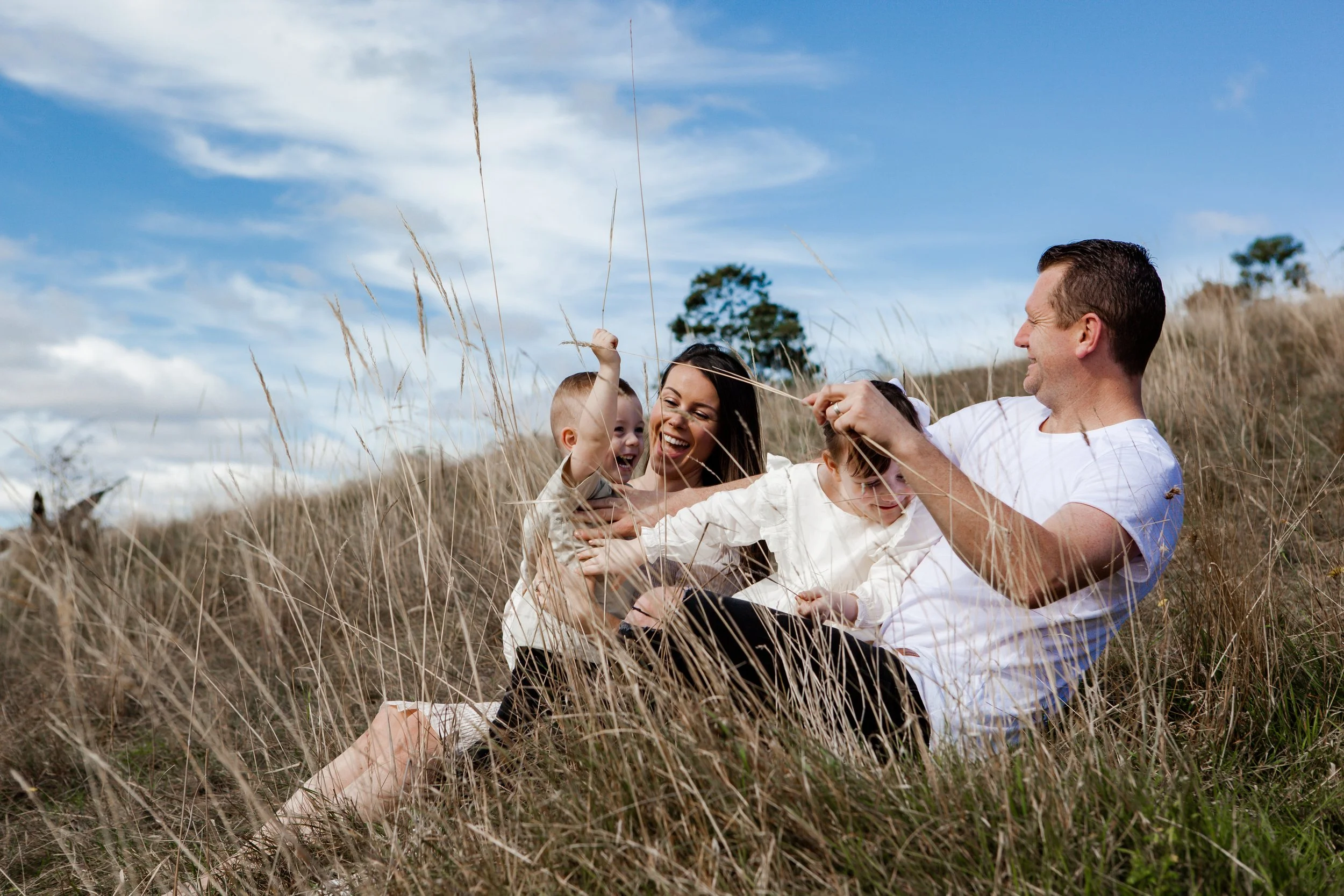Family of four playing and laughing in a grassy field on a sunny day.