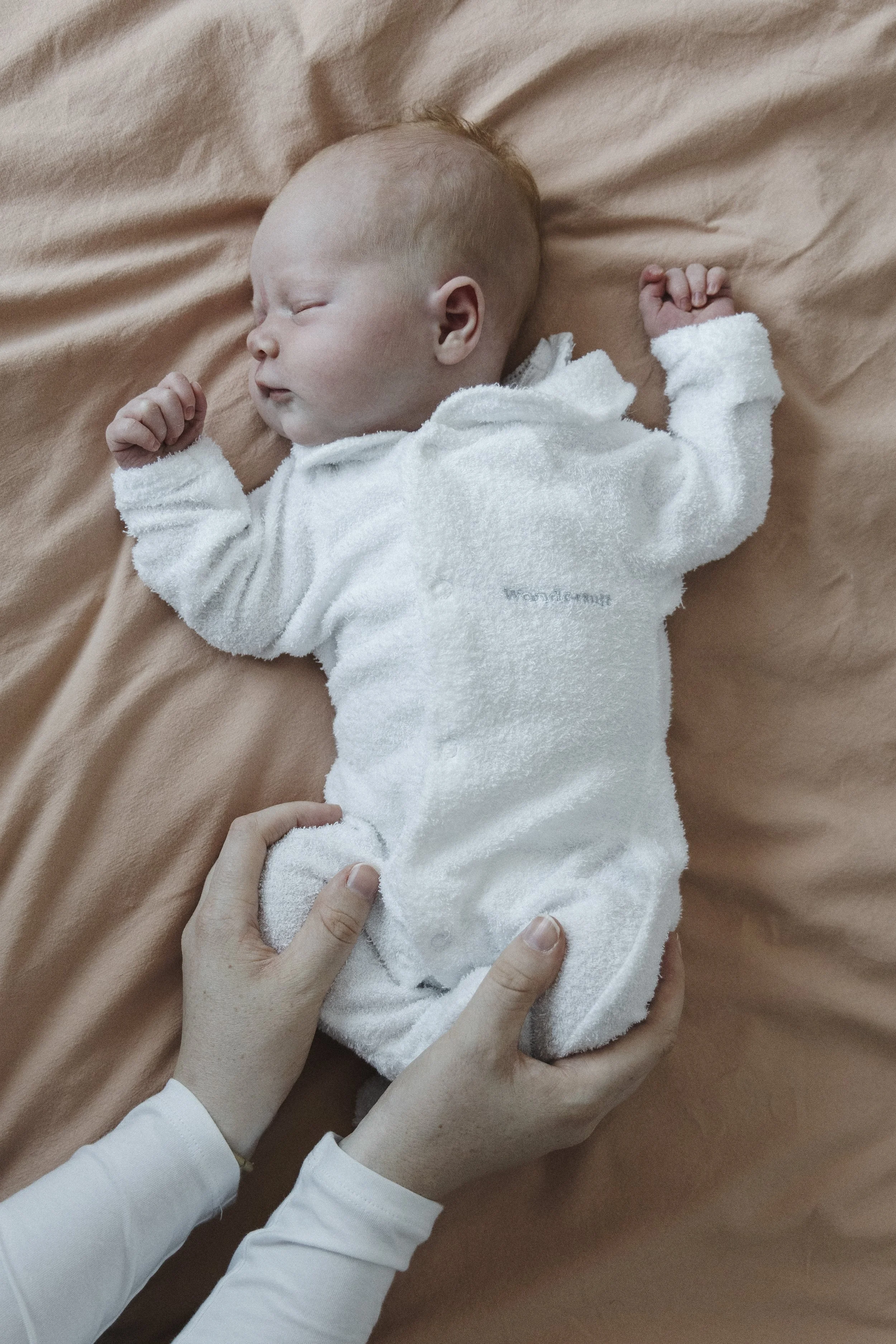 A sleeping baby in a white onesie lying on a light brown blanket, with a person holding the baby's leg.