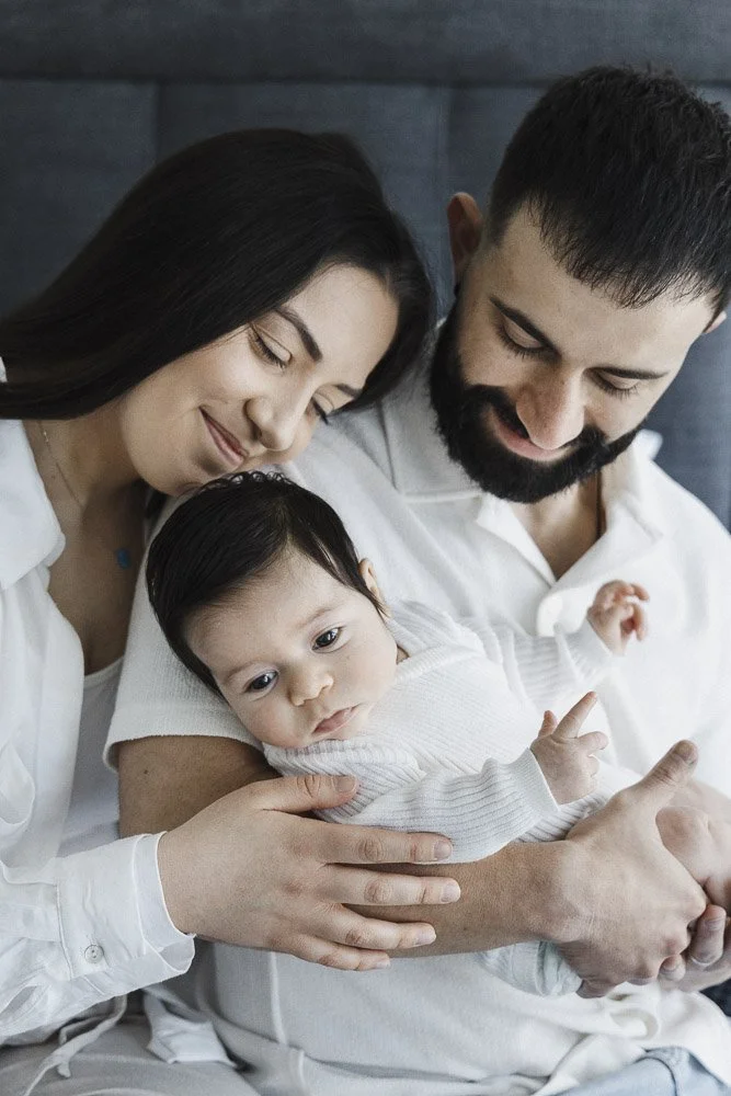 A happy family with a mother, father, and their baby girl sitting together, with the parents smiling at the baby.