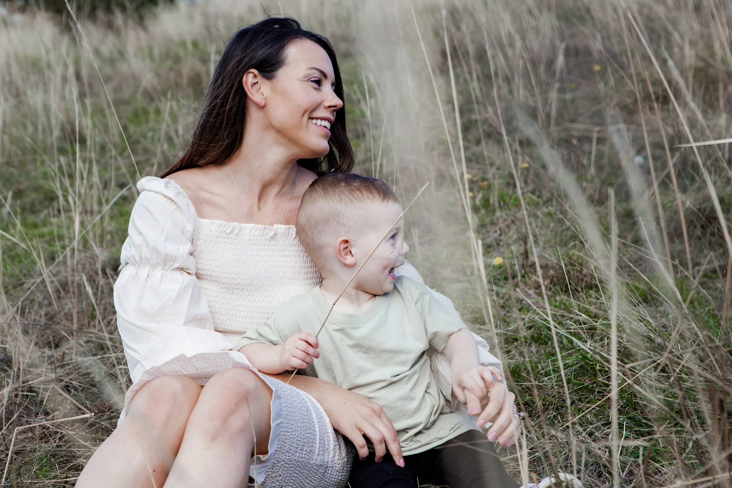 A woman and young boy sitting in a field of dry grass, smiling and enjoying each other's company.