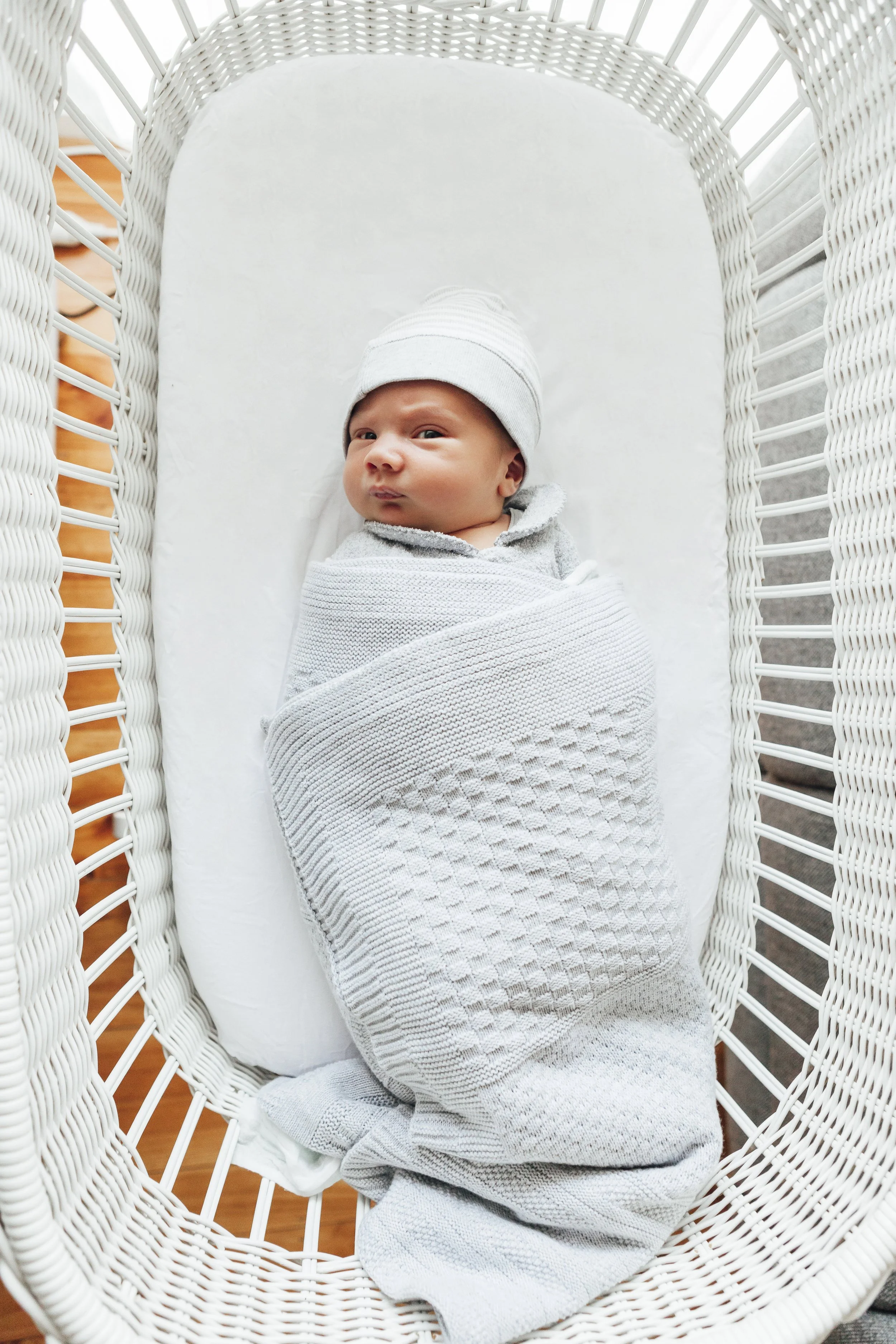 A newborn baby wrapped in a white blanket and wearing a knit hat, lying in a white wicker bassinet.