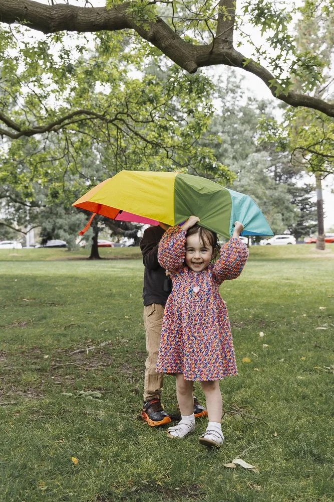 A young girl in a colorful dress and white shoes holding a multicolored umbrella, standing on a grassy park with trees and a row of parked cars in the background.