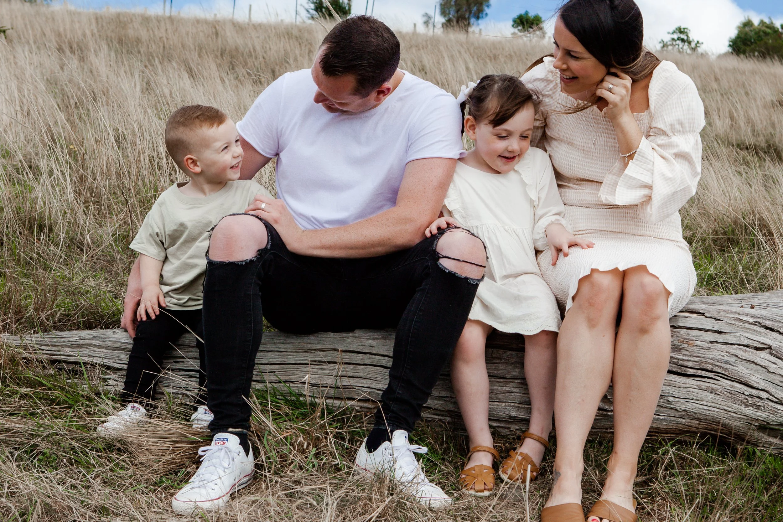 A family of five sitting on a log in a grassy field, smiling and enjoying each other's company.