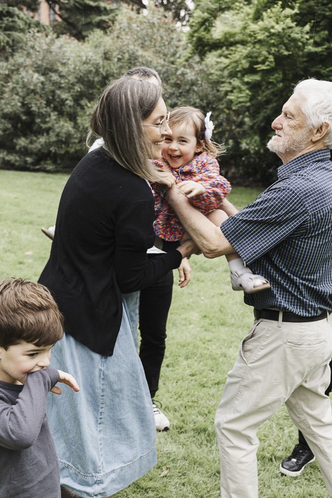 Family outdoors enjoying playful moment, with a woman and an elderly man holding a smiling little girl, and another young boy in the foreground.