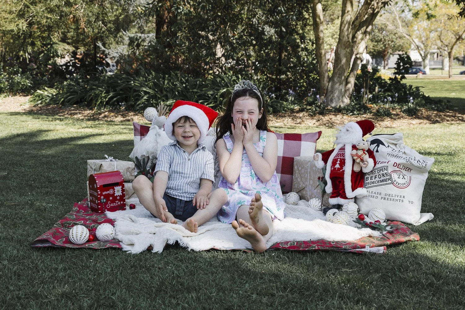 Two children, a young boy wearing a Santa hat and a girl wearing a tiara, are sitting on a blanket outdoors surrounded by Christmas decorations and presents, smiling and laughing.
