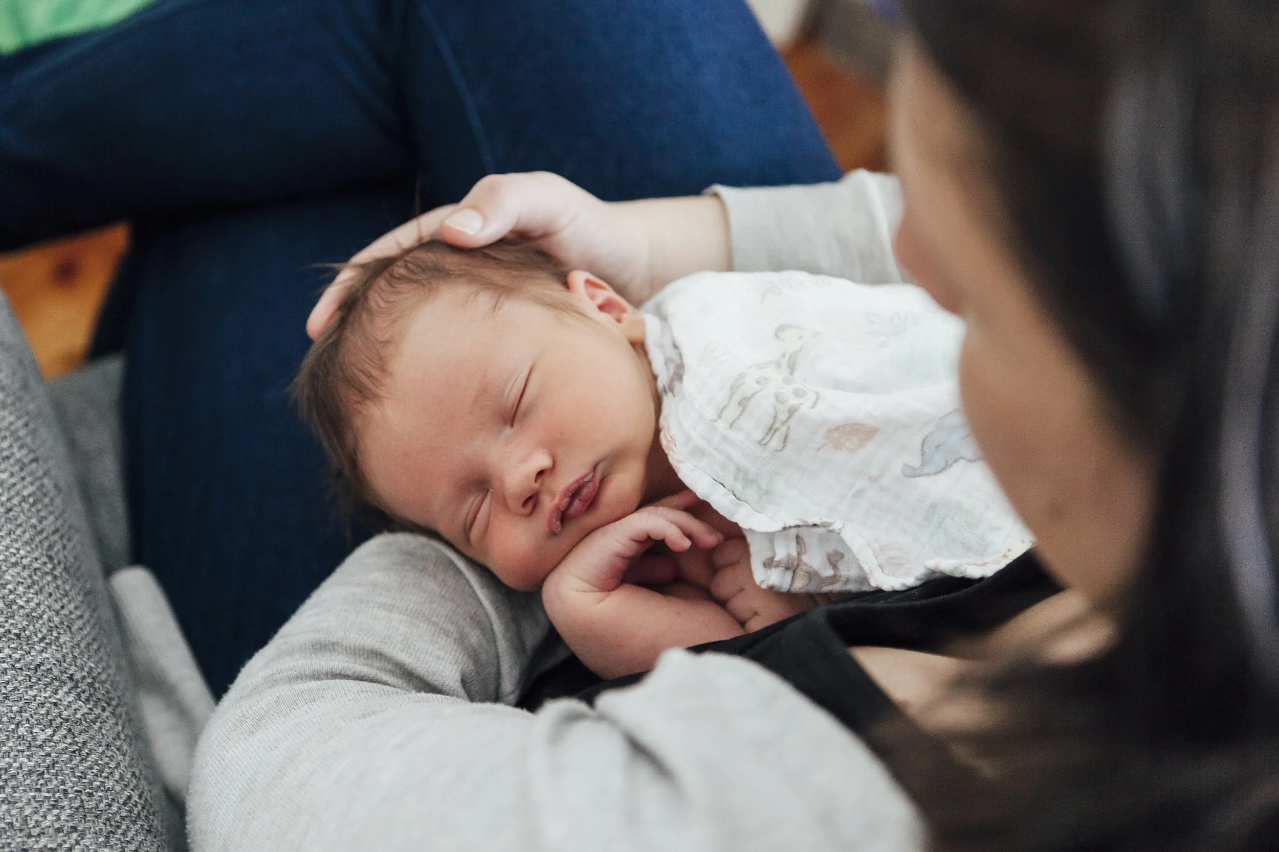 A woman is holding a sleeping baby on her lap, resting her head gently on the baby's head. The baby is wrapped in a white blanket with colorful patterns.