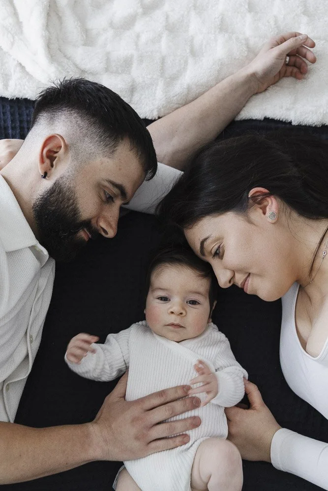 A young family lying on a bed together, with a baby in the center, the father on the left and the mother on the right, all looking at each other affectionately.